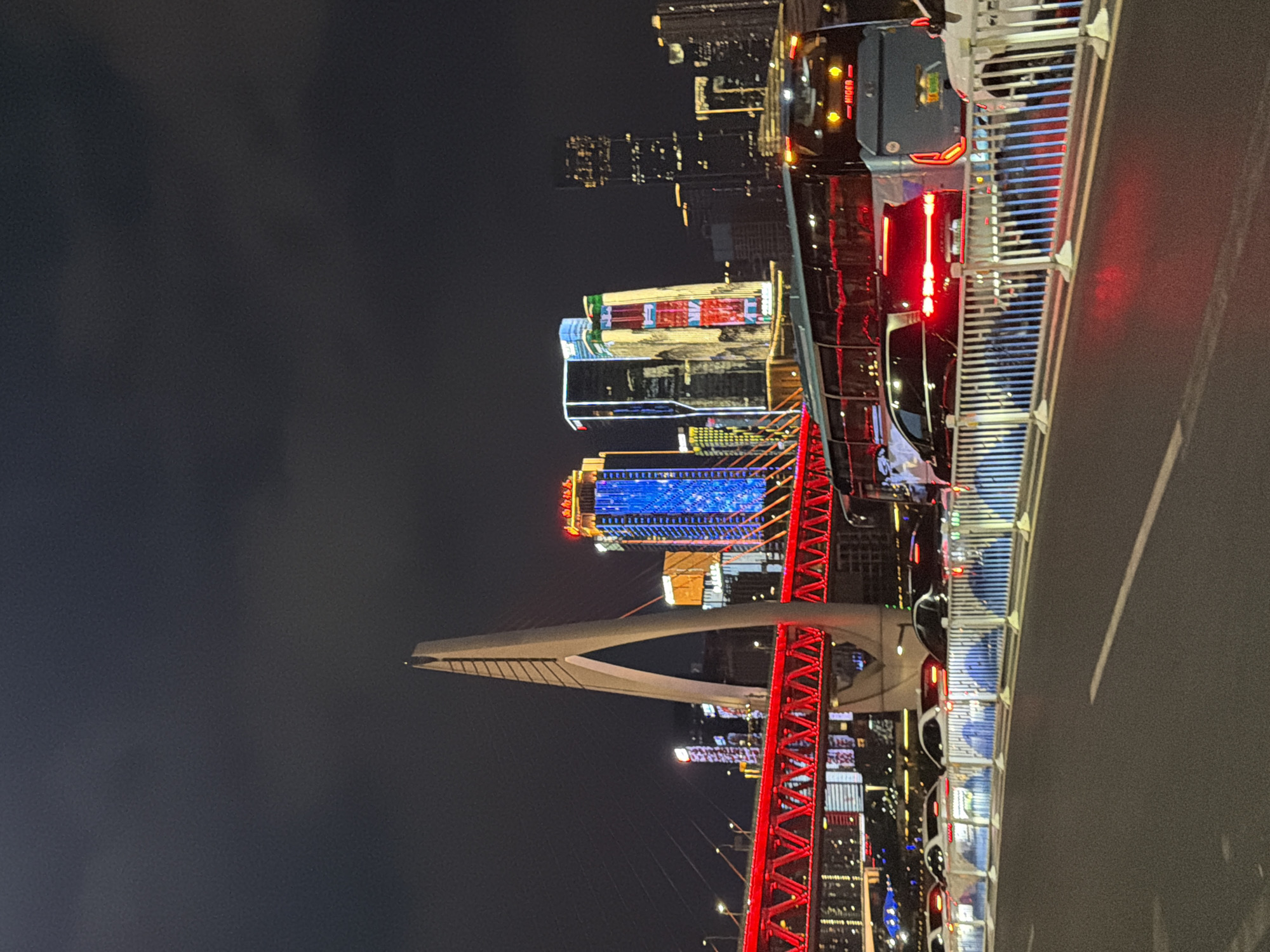 A nighttime view of the city skyline in Yuzhong District, China, features a prominent modern bridge with a tall, white, triangular pylon. The bridge structure itself is illuminated with vibrant red lights. In the background, several skyscrapers are visible, adorned with colorful LED lighting in shades of blue, yellow, and red, creating a lively urban landscape. The scene captures a street-level perspective, with cars and a bus lined up on a road in the foreground, suggesting moderate traffic. The road surface is asphalt, and a white painted line marks a lane. A series of white metal barriers with blue reflective elements are positioned along the side of the road. The sky is dark and overcast, common for nighttime city photography, with no stars or moon visible. The overall impression is one of a bustling, modern metropolis at night, characterized by striking architecture and bright illumination.
