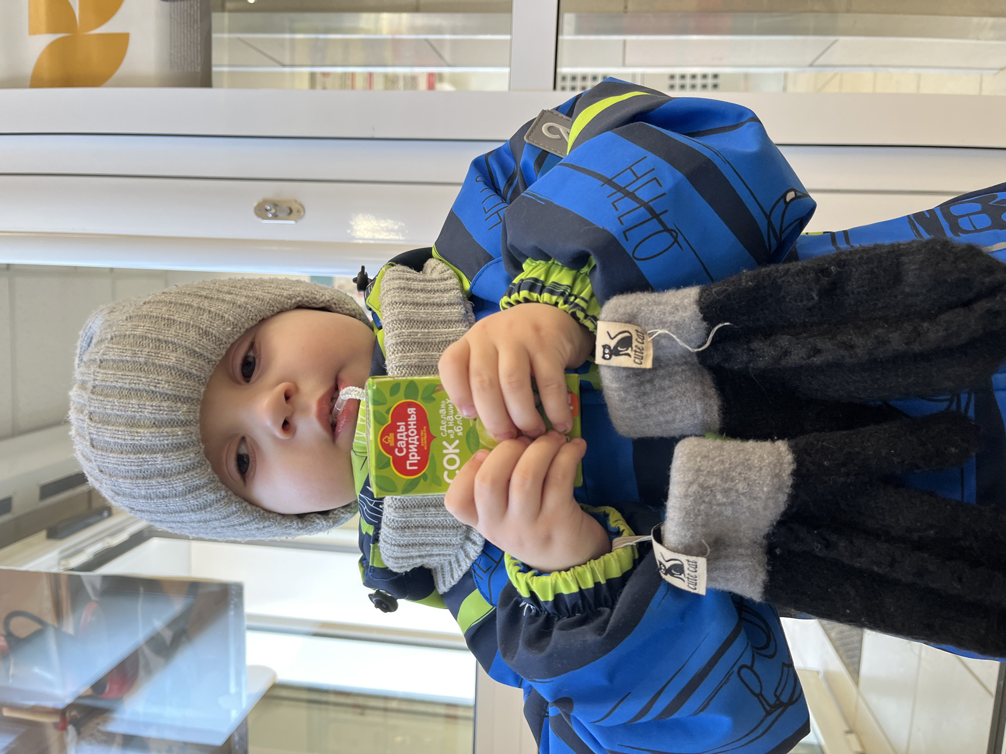 A child in a gray knit hat and a blue and black striped jacket with green trim is holding a green juice box, labeled "Сады Придонья" (Sady Pridonya), with a straw in their mouth. The child is also wearing gray knitted mittens with black cuffs, and one of the mittens has a "cute cat" tag. The location is Chaikovsky, Russia.