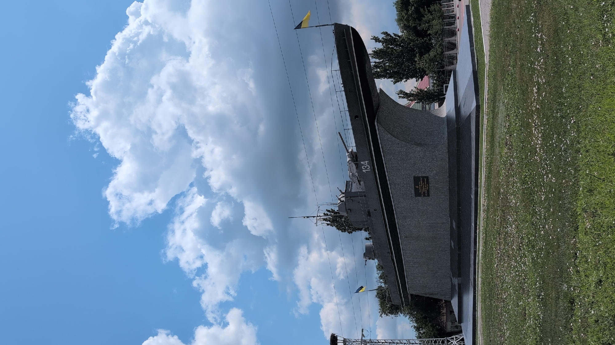 An outdoor, eye-level, medium shot captures a monument of a submarine resting on a stone base, set against a cloudy blue sky and green grass with small white flowers. The submarine, painted dark gray, is positioned at an upward angle, revealing its hull and deck, which features two turrets and a flag pole with a yellow and blue flag. The number "134" is visible on the side of the submarine. A plaque is affixed to the front of the stone base, but its text is illegible. To the left of the monument, a tall, metallic tower with a bird's nest at the top stands prominently, and to the right, a line of green trees and fencing is visible.