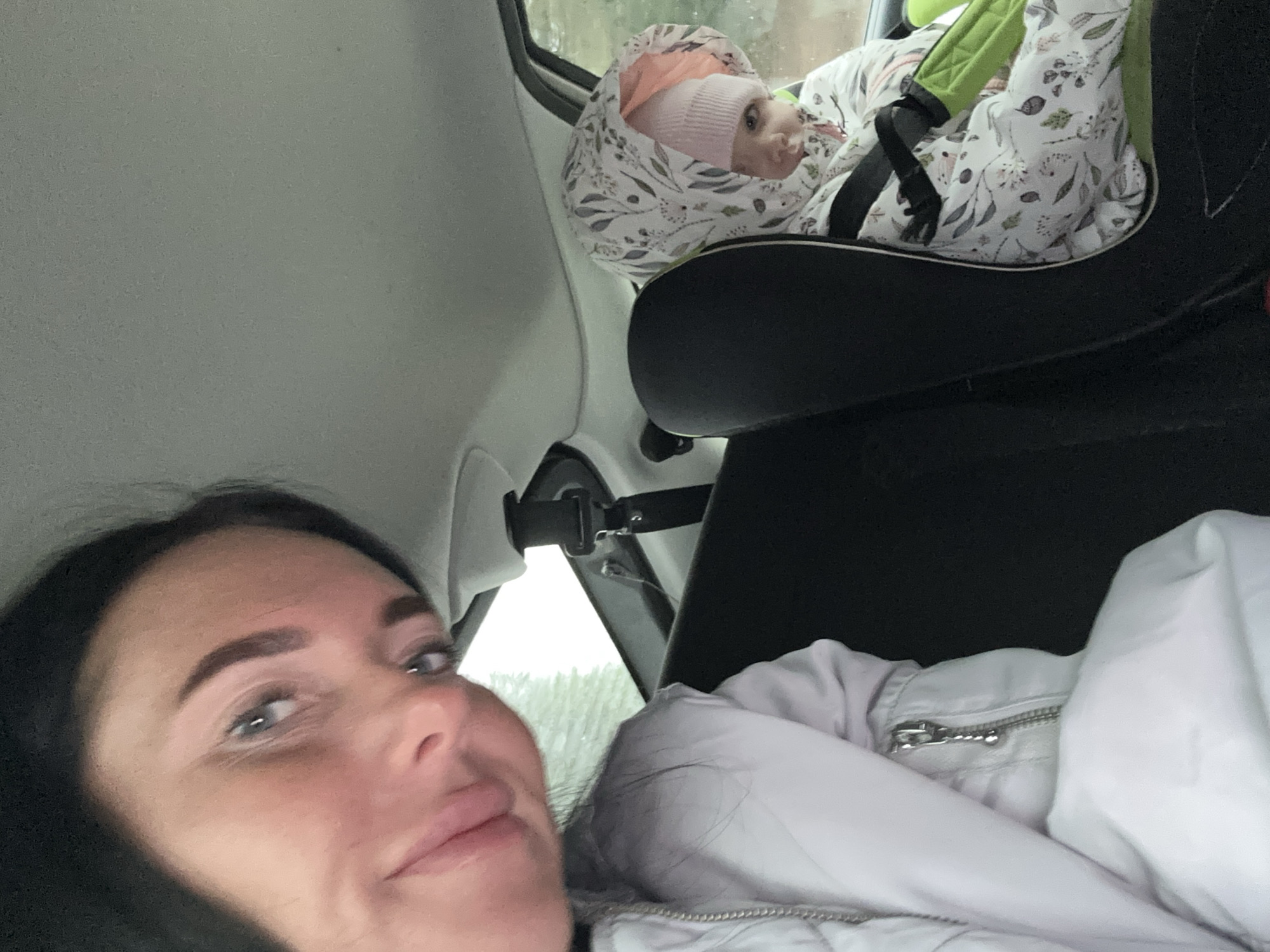 The image shows a woman in the front seat of a car with a baby in a car seat in the back. The woman is fair-skinned with dark hair and is looking at the camera. The baby is wearing a white floral-patterned garment and a pink hat. The location appears to be in a car, as evidenced by the car's interior and a glimpse of trees outside the window. The geographical location of Ольховка, Russia, is not directly identifiable from the visual elements in the image.