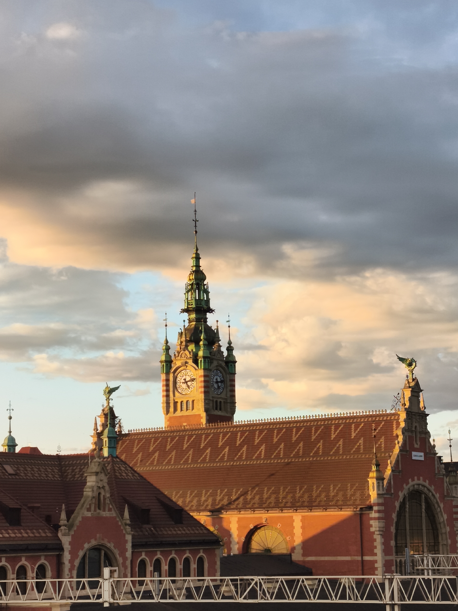 The image shows the Gdańsk Główny train station in Gdańsk, Poland, captured at sunset. The clock tower of the station is prominently featured, with its ornate green spire and clock faces. The terracotta tiled roofs of the station buildings are illuminated by the warm, golden light of the setting sun. Decorative elements, including statues of winged figures on top of some of the building sections, are visible. The sky is filled with dramatic clouds, some tinged with orange and pink hues from the sunset. A white metal structure, likely a railing or bridge, is visible in the foreground. There are no people or vehicles depicted in the image. The scene conveys a sense of historical architecture bathed in the atmospheric light of dusk.