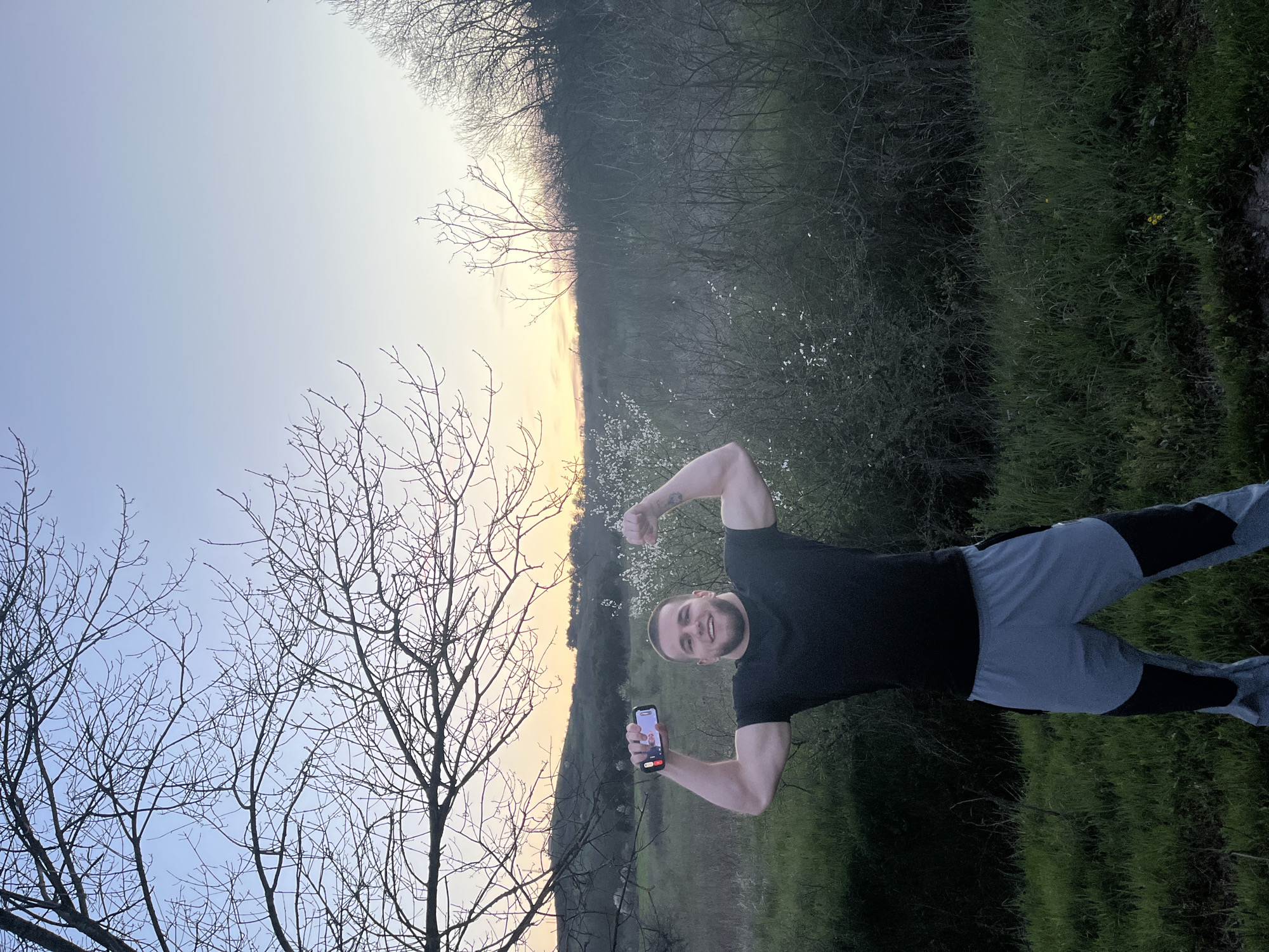 A man stands in a grassy field, holding a phone in his left hand and flexing his right bicep. He is wearing a black t-shirt and gray and black athletic pants. The background consists of a path leading toward a sunset, with trees on either side. It is not possible to determine if this photo was taken in the city of Burgas, Bulgaria with the given information.