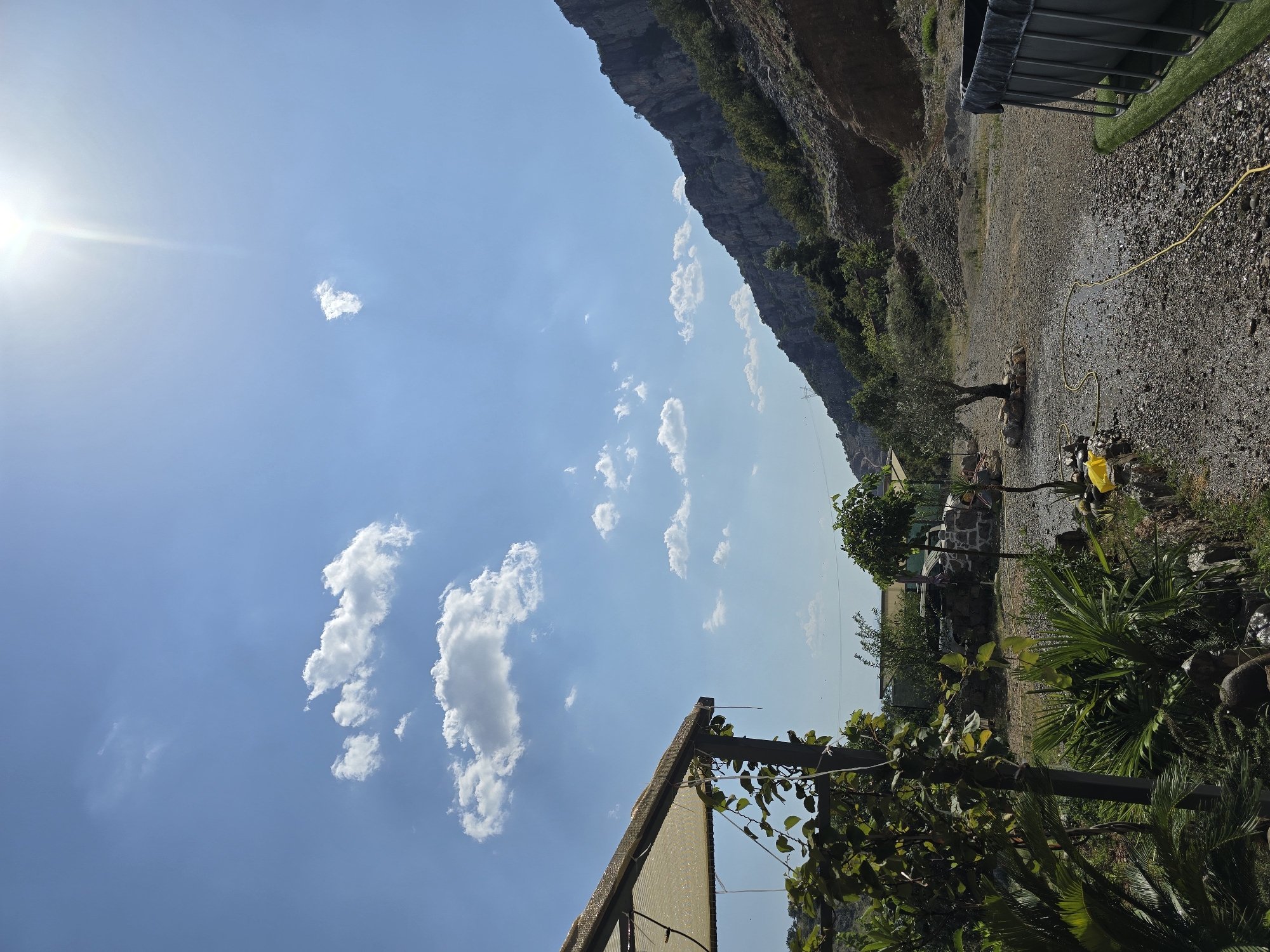 An outdoor scene in a rural or semi-rural area, possibly on the outskirts of Podgorica, Montenegro, is depicted under a bright, sunny sky with scattered white clouds. In the foreground on the left, part of a shaded structure with a tan canvas roof is visible, supported by dark posts and draped with green vines. Lush green vegetation, including palm-like plants and other foliage, occupies the lower left portion of the frame.
The ground is covered with gravel and small stones, with a yellow hose snaking across it. Further into the scene, a small, sparse olive tree stands on a bed of rocks. Behind the tree, there are some low buildings with stone walls and a light-colored awning. The terrain slopes upwards towards a rugged, rocky mountain range that dominates the right side of the image. The mountain is sparsely covered with green shrubs and trees. The sun is high in the sky, casting bright light and creating some glare. On the far right, a portion of a rectangular, dark-colored above-ground pool with a metal frame is visible, situated on a patch of artificial green turf. The overall impression is one of a natural, somewhat undeveloped setting with signs of human activity.