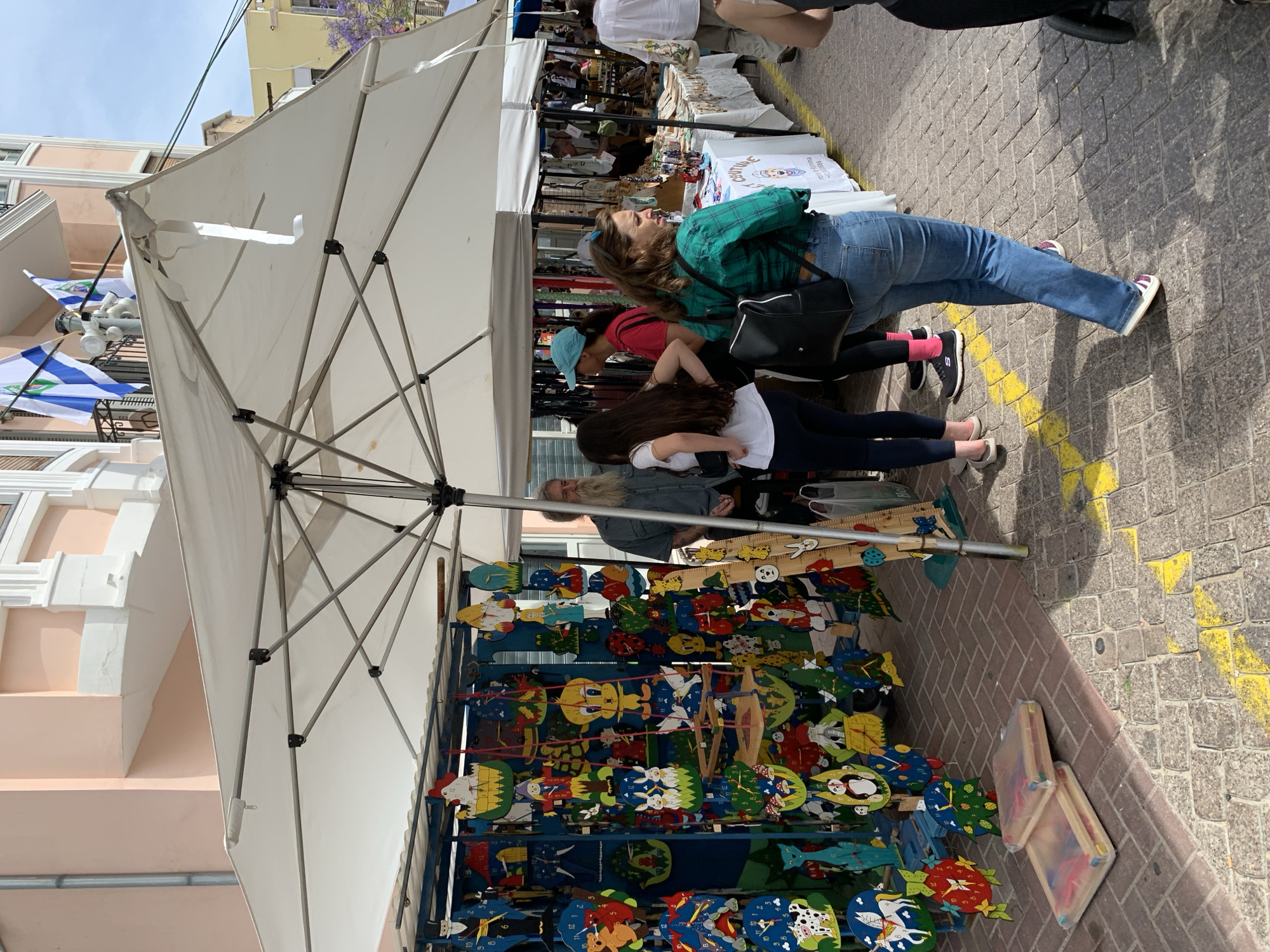 This image shows a street market scene in Tel Aviv, Israel, with a vendor selling wooden clocks and toys. The merchandise is displayed on a multi-tiered metal rack under a large white umbrella. Several people are browsing the items. A woman wearing a green plaid shirt and jeans is looking at the clocks. A younger person in a white shirt and dark pants is standing beside her. An older man with a long beard is also present near the vendor's stall. The market appears to be set up on a brick-paved street marked with yellow paint. Buildings are visible in the background, including one adorned with Israeli flags.