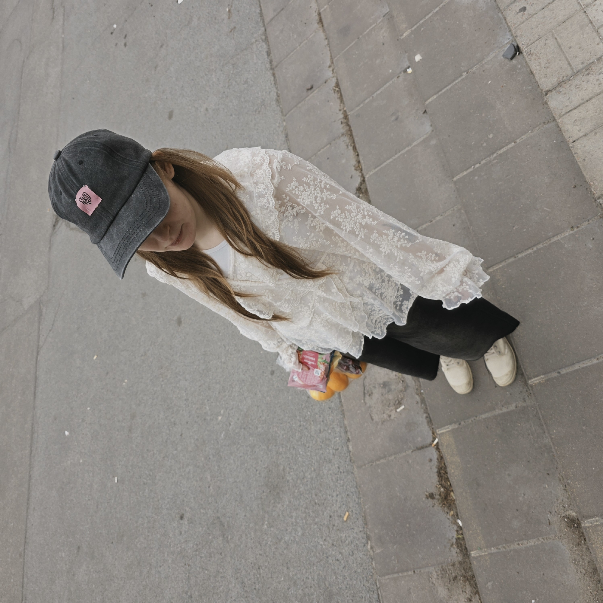 An overhead shot depicts a woman standing on a street, likely in Saint Petersburg, Russia, given the pavement style. She wears a dark baseball cap with a pink patch, a white lace top, black pants, and off-white sneakers. She holds a package of sliced meat and oranges. The background includes asphalt and tiled sidewalk. Cigarette butts lie scattered on the ground.