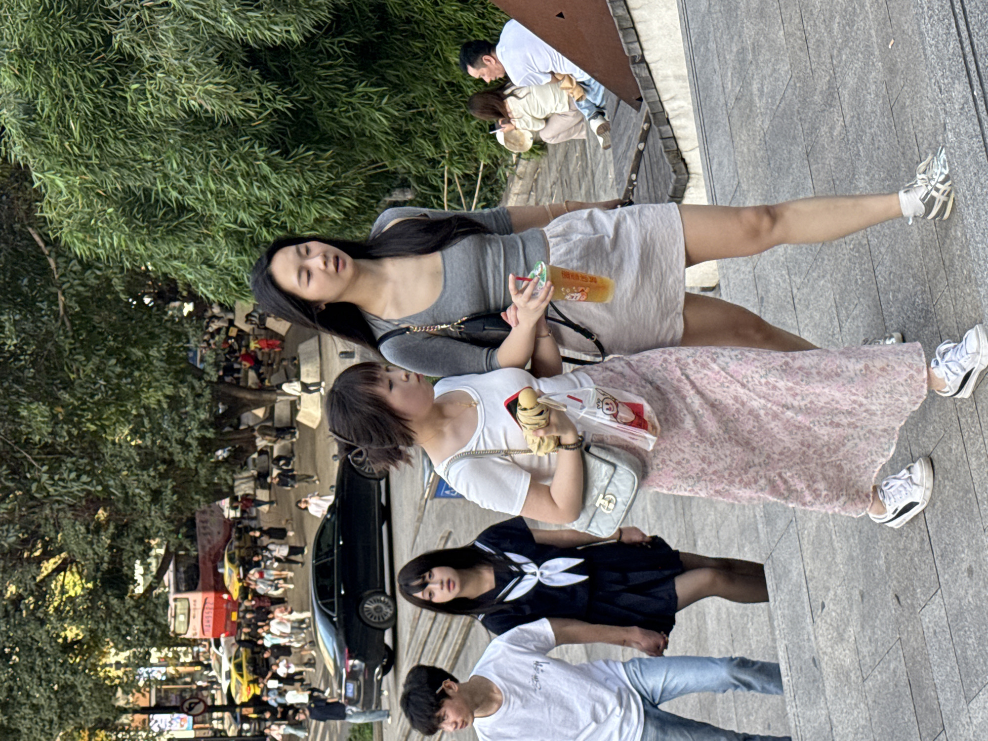In the Yuzhong District of China, a street scene presents a view of pedestrians walking on a paved area beside a road with vehicles. The road is lined with shops and a bus. The crowd on the street includes numerous individuals, creating a bustling atmosphere. Foregrounded, a young woman in a grey top and a light skirt holds a beverage. Next to her, a second woman wearing a white top and a floral skirt carries a bag. The bottom foreground features a woman in a navy dress and a man in a white t-shirt, both walking. Foliage and trees are present on the left side of the frame. In the background, more people can be seen sitting and standing. The entire composition captures a normal day with people going about their lives in an urban environment.