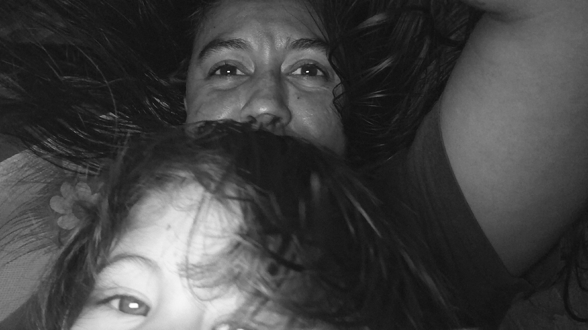 This black and white image, captured in Vallecito, Venezuela, portrays two individuals in a close-up, diagonal composition. An adult female, positioned on the left side of the frame, is depicted lying down with dark, wavy hair fanned out around her face. Her eyes are open and directed towards the viewer, and facial features including her nose and mouth are visible. A dark garment is partially visible on her upper body. Adjacent to her on the right is a young child, also lying down, whose face is partially obscured. The child's right eye is clearly visible, gazing directly towards the viewer, and some lighter-toned hair is spread around their head. A small, light-colored, multi-petaled flower accessory is visible within the child's hair near the right ear. The background is dark and featureless, indicative of an indoor setting.