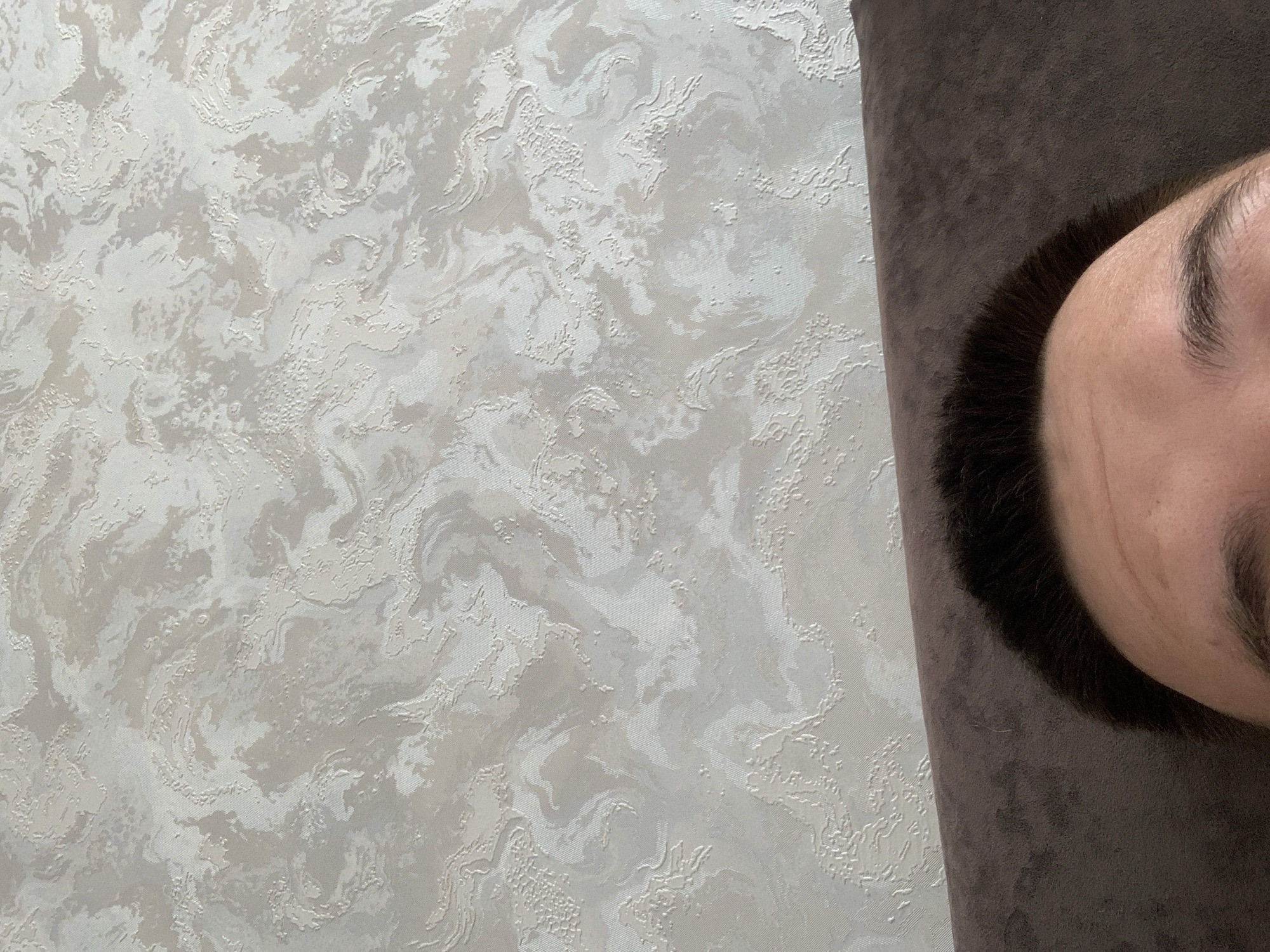 The image shows the upper portion of a person's face and their dark hair, with their forehead and eyebrows visible. The person is lying down, with their head resting on a dark brown, textured surface, likely a sofa or bed. Behind the person is a wall with a textured wallpaper featuring a swirling, marbled pattern in light beige and off-white tones. The image is framed from a low angle, looking upwards. There is no indication of the city of Stavropol, Russia, within the visual information provided in the image.