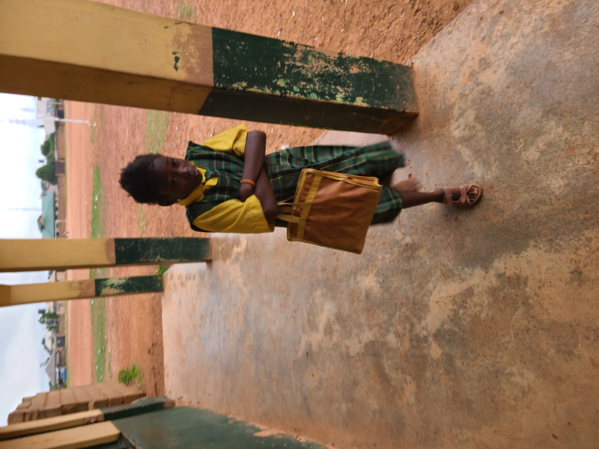 A young person, likely a child, stands under a covered walkway in Rafin Bauna, Nigeria. The child, positioned slightly to the right of the frame, looks directly at the camera with a neutral expression. They are dressed in a school uniform, featuring a short-sleeved yellow top and a green and black plaid dress or pinafore. A light brown bracelet adorns their right wrist, and they wear light-colored sandals on their feet.
The child holds a rectangular, light brown or tan bag with darker brown straps, possibly a school bag. They are standing on a worn, discolored concrete floor. To their left and right are sturdy pillars, painted in alternating bands of yellow and dark green, showing noticeable wear and chipped paint. These pillars and the concrete floor suggest a school building or a similar community structure.
In the background, beyond the covered area, lies a wide expanse of reddish-brown dirt, with sparse green vegetation scattered across it. Distant, simple buildings and what appears to be a mosque minaret are visible under an overcast sky. The lighting is soft and diffuse, indicating daytime under cloud cover, with no harsh shadows. The scene evokes a sense of daily life in a school or community setting in a semi-rural Nigerian environment. No visible text is present in the image.