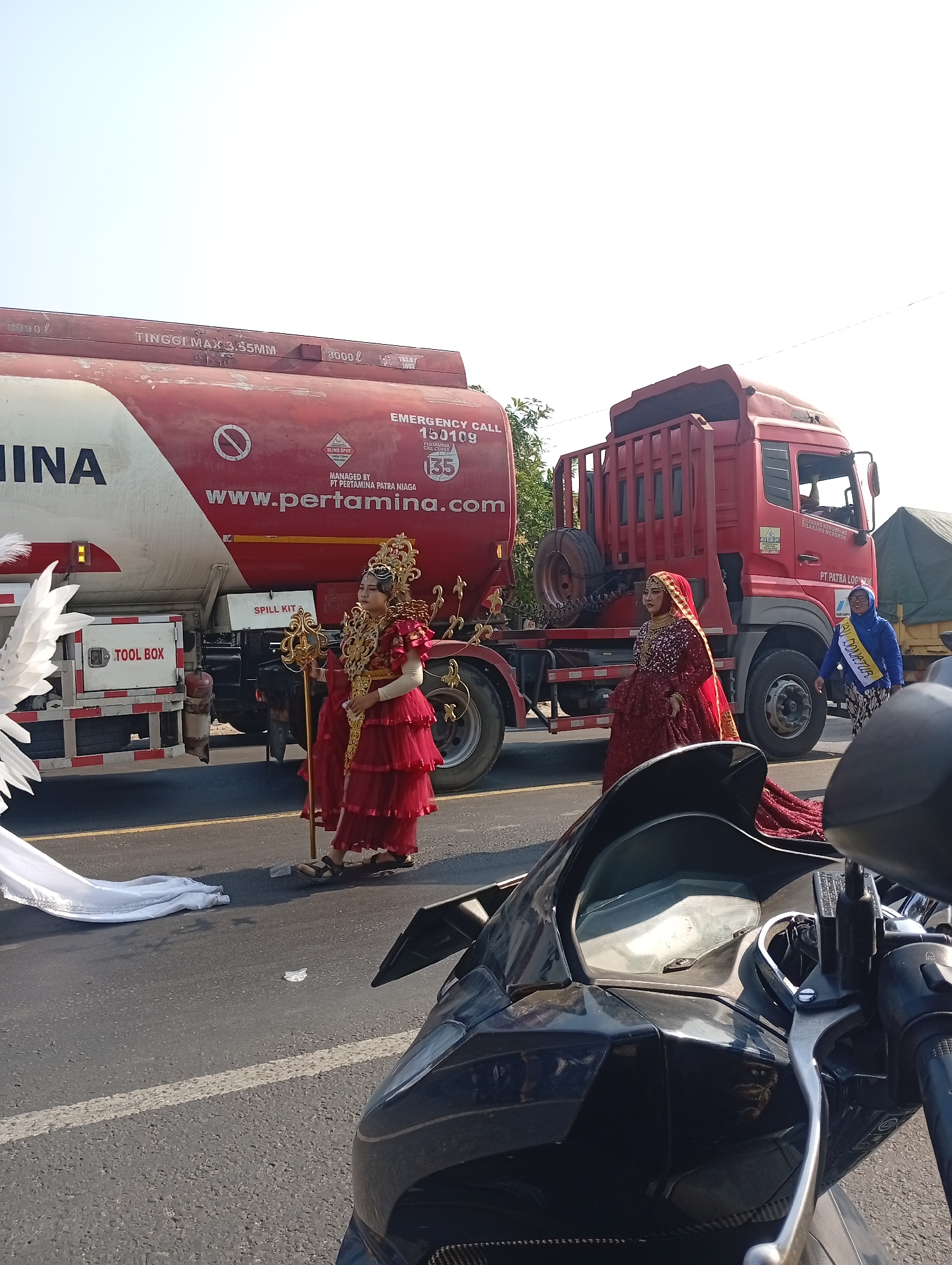 This image depicts a street scene in Sarang, Indonesia, possibly during a parade or event. Two women in elaborate, traditional attire are standing on the asphalt road next to a large red Pertamina fuel truck. The woman on the left wears a red tiered dress with gold adornments and a tall, ornate headdress, holding a golden staff. The woman on the right is dressed in a shimmering red gown and a matching headscarf, appearing to be Indian-inspired attire. A third woman in a blue headscarf and a yellow sash with text partially visible is standing further back near another truck. The fuel truck has "PERTAMINA" branding prominently displayed, along with an "EMERGENCY CALL 150109" notice. To the left of the fuel truck, a white fabric, possibly part of a costume or decoration, lies on the ground. In the foreground, the handlebars and part of the dashboard of a dark-colored motorcycle are visible, suggesting the photo was taken from a rider's perspective. The sky is clear and bright, indicating a sunny day.