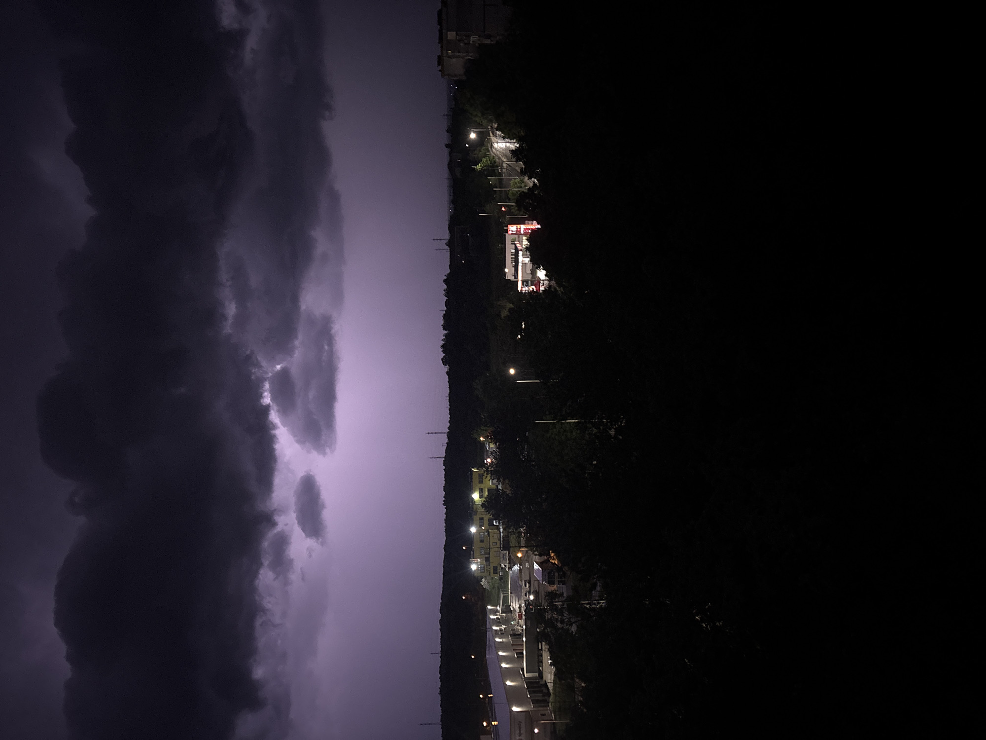 The image depicts a night scene in Pleven, Bulgaria, dominated by a dramatic lightning storm. The left side of the frame is filled with dark, ominous storm clouds illuminated by the light of the lightning. The right side shows a cityscape under the storm, with building lights visible through the darkness. The area appears to be partially wooded, with tree lines separating buildings. In the distance, the identifiable shape of a red and white structure can be seen, possibly a commercial building or complex. Streetlights dot the area, adding to the overall illumination. The foreground is mostly dark, obscuring details close to the camera. The scene suggests a moment of active weather affecting the urban landscape.
