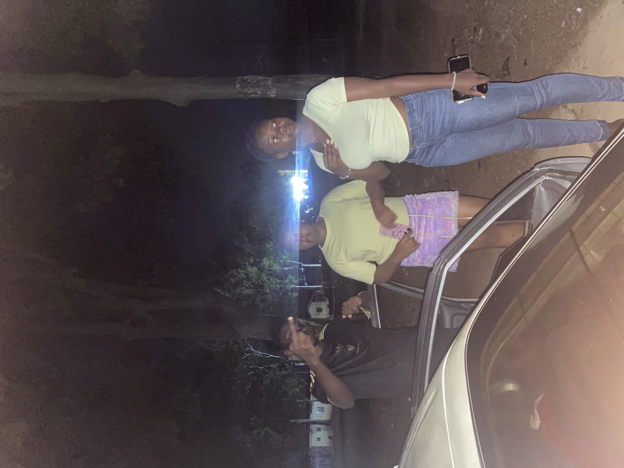 The image, taken at night in Rafin Par, Nigeria, features three young people standing near a car. The first subject, a young woman, is leaning against a car with her right arm on the vehicle and her left hand holding a smartphone. She is wearing a light yellow crop top and blue jeans. The second subject, also a young woman, is standing to her left and appears to be looking down at her phone. She is wearing a light yellow t-shirt and a purple skirt. The third subject, a young man, is standing in front of the second subject and is giving a middle finger gesture towards the camera. He is wearing a black t-shirt with yellow stripes. The background consists of trees, a dirt road, and some small buildings with lights on, suggesting a rural or semi-urban setting. The presence of artificial lights in the background and the overall darkness indicate that the photo was taken after sunset. The atmosphere appears casual and social, with the subjects interacting with each other and the camera. There is no visible text in the image.