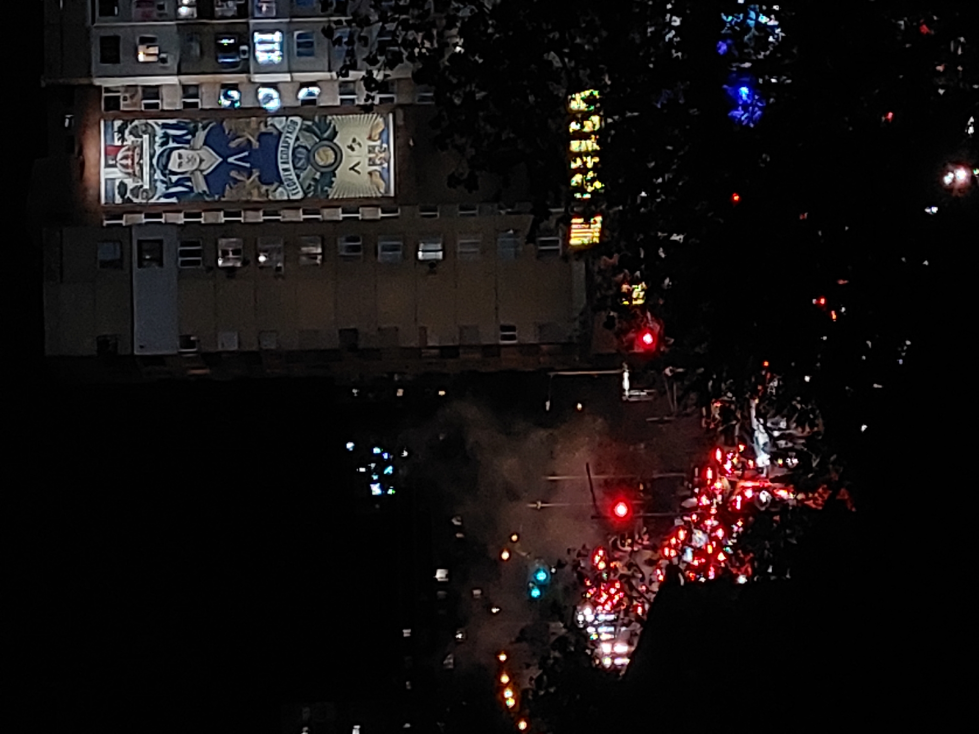 Captured at night, the scene presents an overview of a cityscape in an unknown location. The buildings and street are illuminated by artificial lights, with traffic signals and vehicle headlights adding to the luminance. The dominant structure in the frame is a large building adorned with a mural of a person in uniform, alongside an emblem or crest. The scene also captures what appears to be an illuminated vertical sign. The street below the buildings show traffic and scattered pedestrians. The environment is overall dark, with the available lights painting the scene. The perspective suggests the photo was taken from an elevated position.