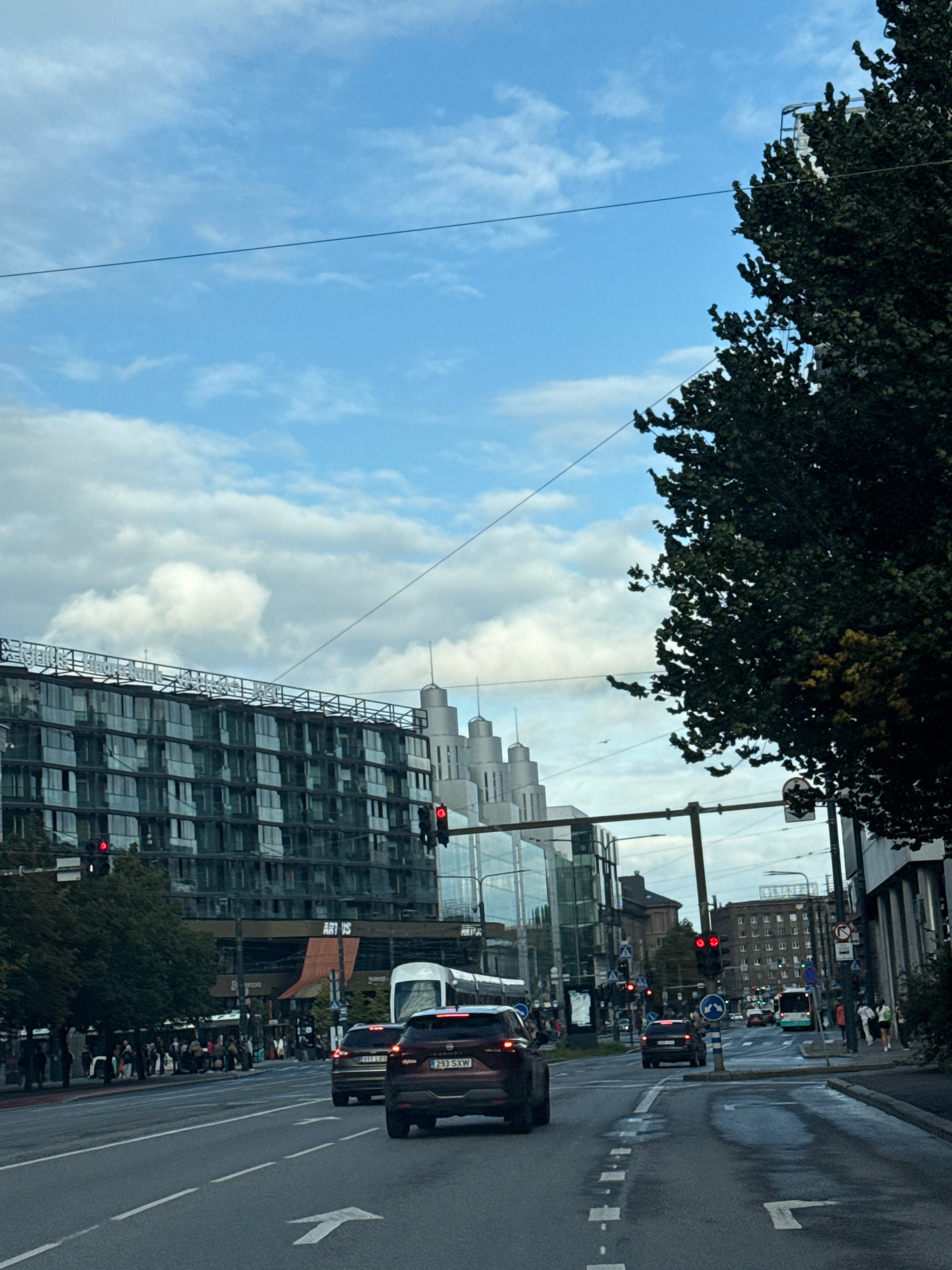 The image captures a view of the city of Tallinn, Estonia, on a cloudy day. A multi-lane road dominates the foreground with vehicles in motion, including two cars and a bus. The road surface shows signs of wetness and reflections. Traffic lights are visible and appear to be red, indicating a stop.
On the left side of the road, a large, modern building with a glass facade and multiple floors stands prominently. Trees line the street in front of this building. Pedestrians are walking on the sidewalk.
Further down the road, other buildings are visible, including a structure with distinctive cylindrical towers. A public transport bus is also seen on the road. The sky is a mix of blue and white clouds, with power lines crisscrossing the upper portion of the frame. The overall scene depicts a busy urban environment.