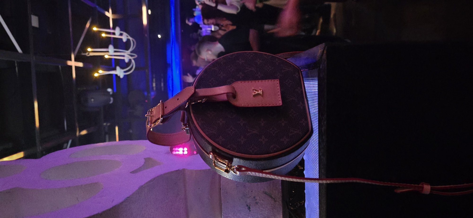 The image shows a Louis Vuitton cylinder purse sitting on a white and pink surface. In the background, there is a crowd of blurred people, and a black metal structure with decorative lights that resemble candelabras. A tall blue light is visible as well. The location is most likely indoors at an event or venue, but the city of Sofia, Bulgaria is not discernible.