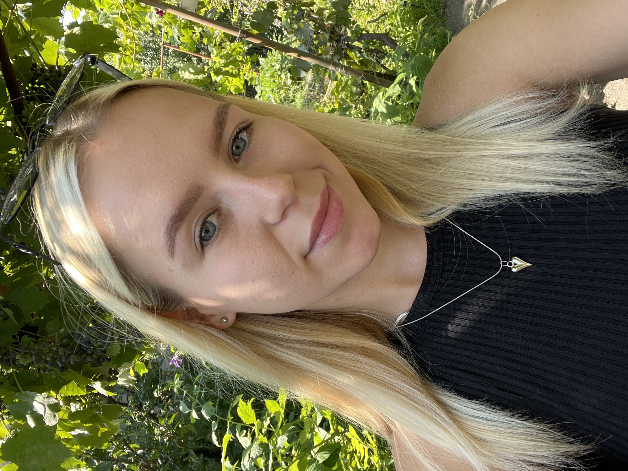A young woman with blonde hair and blue eyes is taking a selfie outdoors. She is wearing a black sleeveless shirt and a silver heart-shaped pendant necklace. Behind her, there are green leaves and bunches of grapes on a vine. The lighting suggests it is a sunny day.
