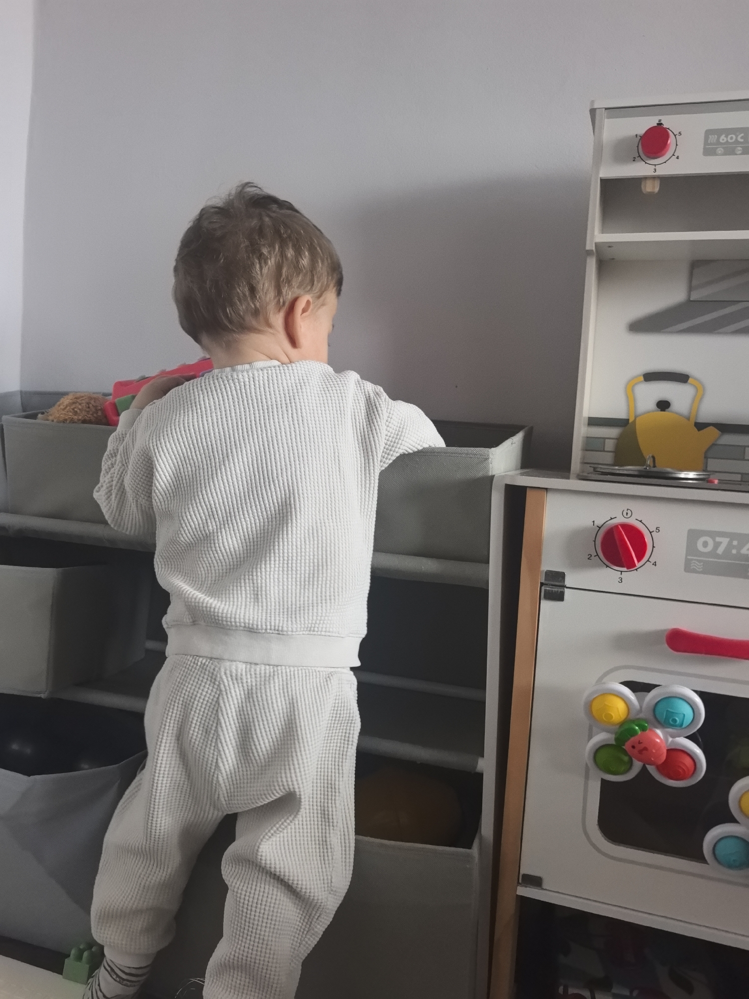 An indoor domestic scene is depicted, identified as being in Stara Zagora, Bulgaria, though no specific address or landmark is visible. A young child, viewed from the rear, is the primary subject. The child has light brown hair and wears a matching light-colored, waffle-knit long-sleeved shirt and pants. The child's left foot, partially visible, shows striped socks. The child is actively leaning into or reaching towards the top-most grey fabric storage bin of a multi-tiered shelving unit positioned against a plain, light grey wall. Inside this bin, a brown plush toy and a red object are discernible. To the child's right, a white wooden toy kitchen unit is present. The top section of the toy kitchen features a red circular dial set to the number '5' and text indicating "60°C". Below this, a graphic depicts a yellow kettle on a stovetop with a tiled backsplash pattern. The lower part of the toy kitchen includes another red circular dial set between '1' and '2', a digital display showing "07:4", and a red handle. The oven door features multiple colorful spinner toys, including one shaped like a strawberry. The floor is partially visible beneath the child.