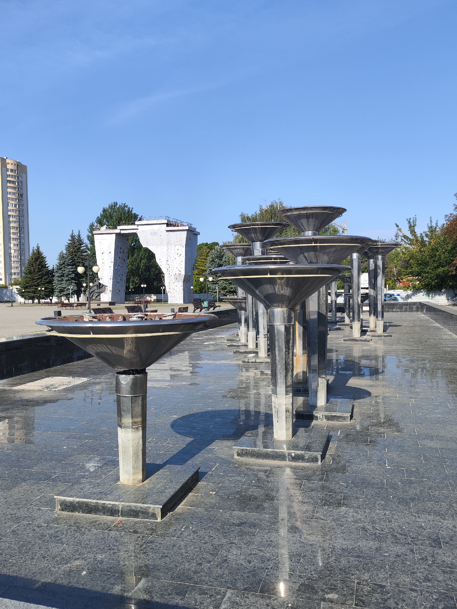 The image shows an outdoor public space with a dry fountain in the foreground. The fountain consists of multiple tiered, cone-shaped metallic structures on cylindrical bases, arranged in a linear fashion. The ground is paved with dark granite tiles, some of which are wet, reflecting the structures and the sky. In the background, a tall, light-colored apartment building stands to the left, and a white, arch-shaped structure with climbing holds, likely a climbing wall, is visible behind the fountain. Several evergreen trees and park benches are also present. The sky is clear and blue. This scene is located in Kharkiv, Ukraine, specifically at the area of the "Climbing Wall" fountain in Shevchenko Garden. There are no people visible in the image.