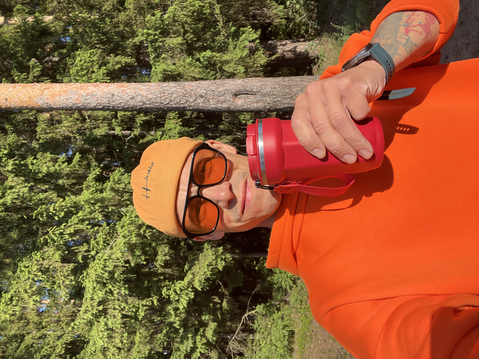 The image captures a male-presenting individual in a vibrant green coniferous forest under bright sunlight, likely in Chaikovsky, Russia. The photo is rotated 90 degrees clockwise.
The main subject is a person wearing an orange hooded sweatshirt and an orange beanie, along with orange-tinted sunglasses featuring black frames. He has light stubble and looks directly towards the camera, suggesting a selfie or a close-up portrait. He holds a red insulated water bottle in his right hand. On his right forearm, a colorful tattoo with red/pink and black elements is visible, along with a dark-colored watch featuring a ribbed gray strap. A small, indistinct embroidered text or logo can be seen on the front of his beanie.
The background is dominated by dense green pine or spruce trees, indicating a natural, wooded environment. A prominent tree trunk, light brown with textured bark, runs horizontally across the mid-ground. The bright lighting suggests it is daytime, casting clear shadows and illuminating the scene vividly. The overall impression is of someone enjoying an outdoor activity, taking a break for hydration in a sunny forest setting.