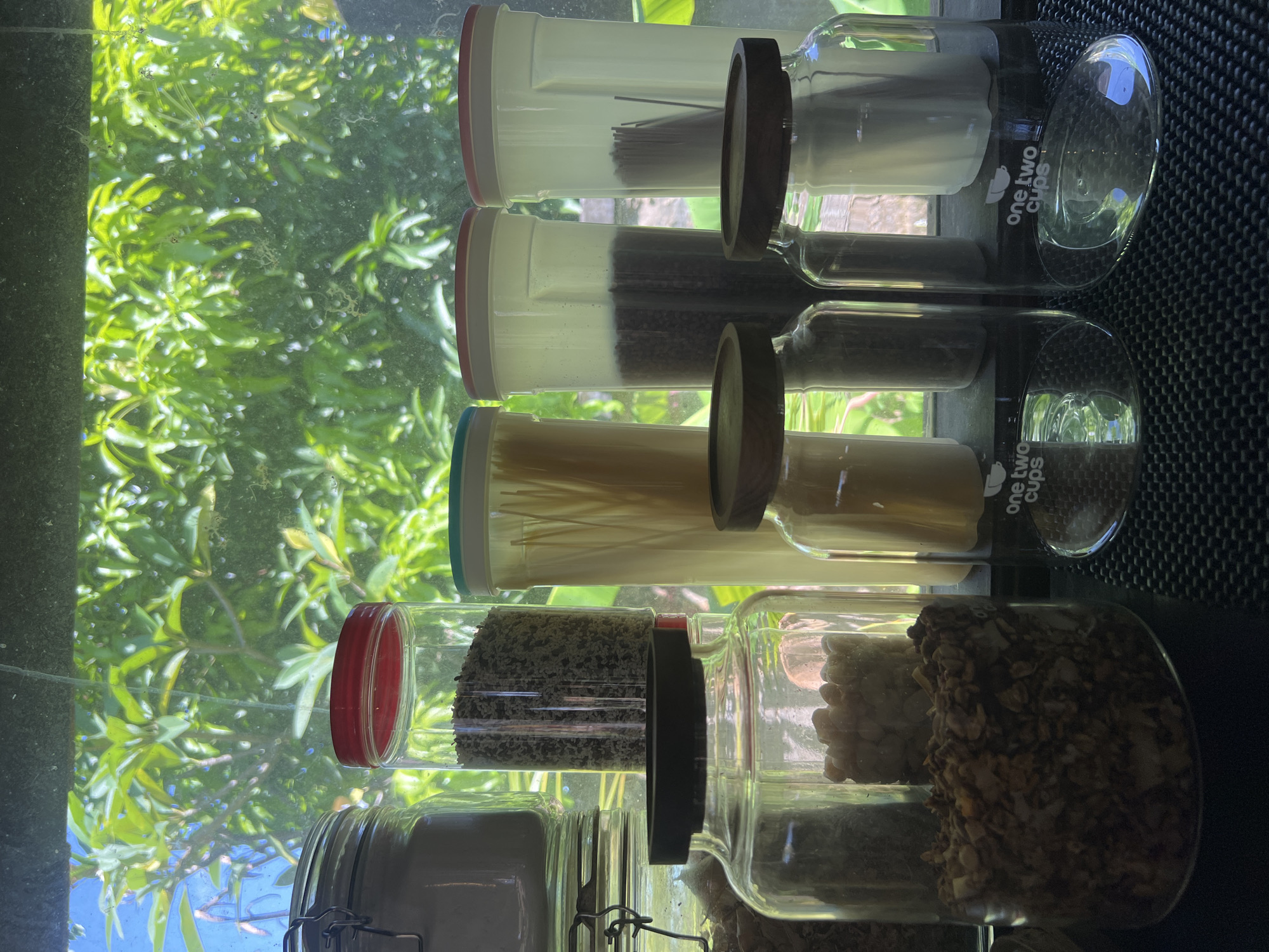The image shows several glass jars with colorful lids stacked vertically against a window. The jars appear to contain dry food items, including pasta, beans, and granola. The window offers a view of lush green foliage, which is likely located in Sukawati, Indonesia. The jars are on a dark countertop. One of the jars has the words "one two cups" printed on it.