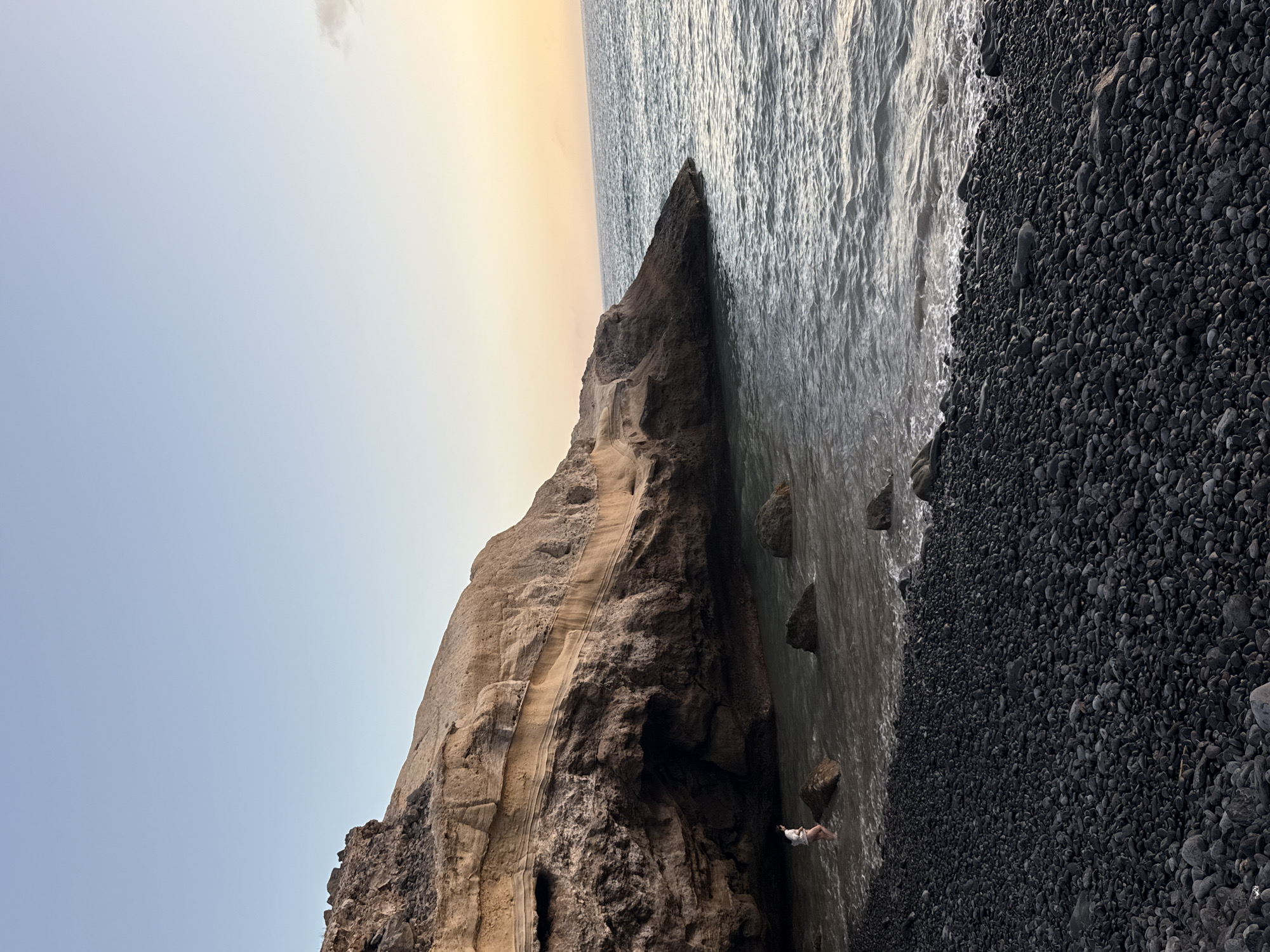 The image captures a serene coastal scene at dusk, likely in Adeje, Spain. The foreground is dominated by a black pebble beach, the stones smooth and varied in size. Gentle waves lap at the shore, creating a soft, frothy edge. In the mid-ground, a person, seen from behind, stands in the shallow water, partially submerged, facing a large, imposing cliff face. The cliff is layered, revealing strata of light brown and darker rock, with a dark cave-like opening near the waterline. Several large, dark rocks are scattered in the water around the person and the cliff. The background showcases the ocean, its surface rippling under the fading light of the setting sun, which casts a warm, golden hue across the horizon. The sky above is a clear, soft gradient of pale blue, transitioning to the warm colors of the sunset. The overall atmosphere is tranquil and natural.