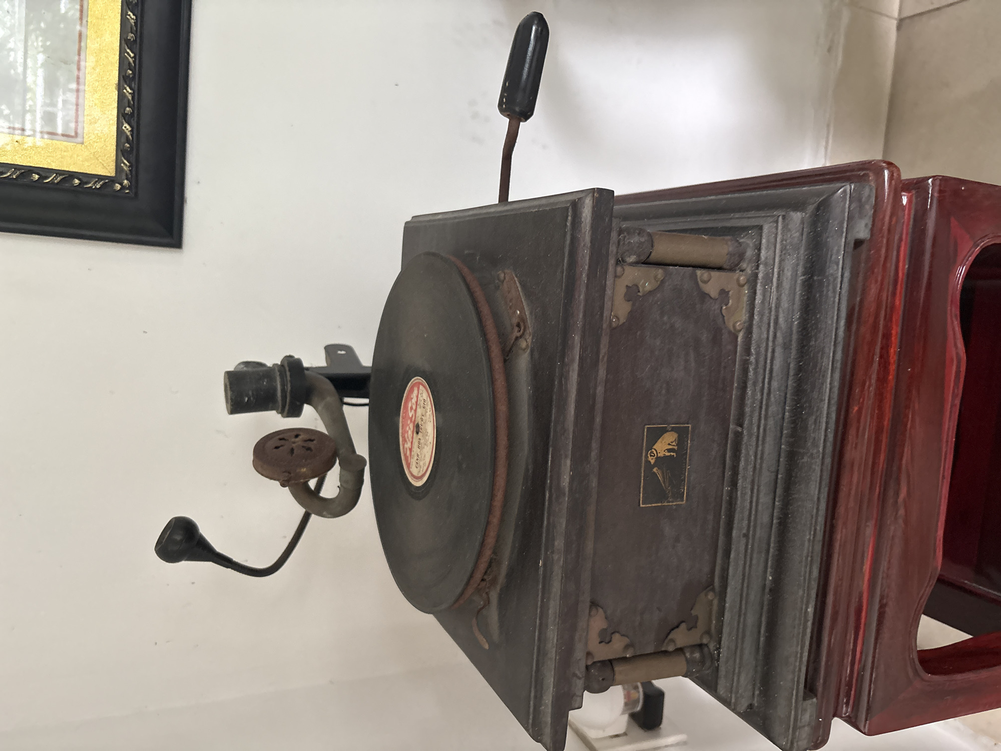 An antique gramophone sits on a dark red wooden stand in a room with white walls. The gramophone is dark brown and has a circular turntable with a record on it. There is a metal arm with a horn attached to it, and a crank on the side of the gramophone. A small emblem with a dog and a horn is visible on the front of the gramophone. The stand is ornate with carved details. A framed picture with a gold border hangs on the wall in the background. The scene is lit by natural light, casting shadows. The overall impression is one of historical preservation.