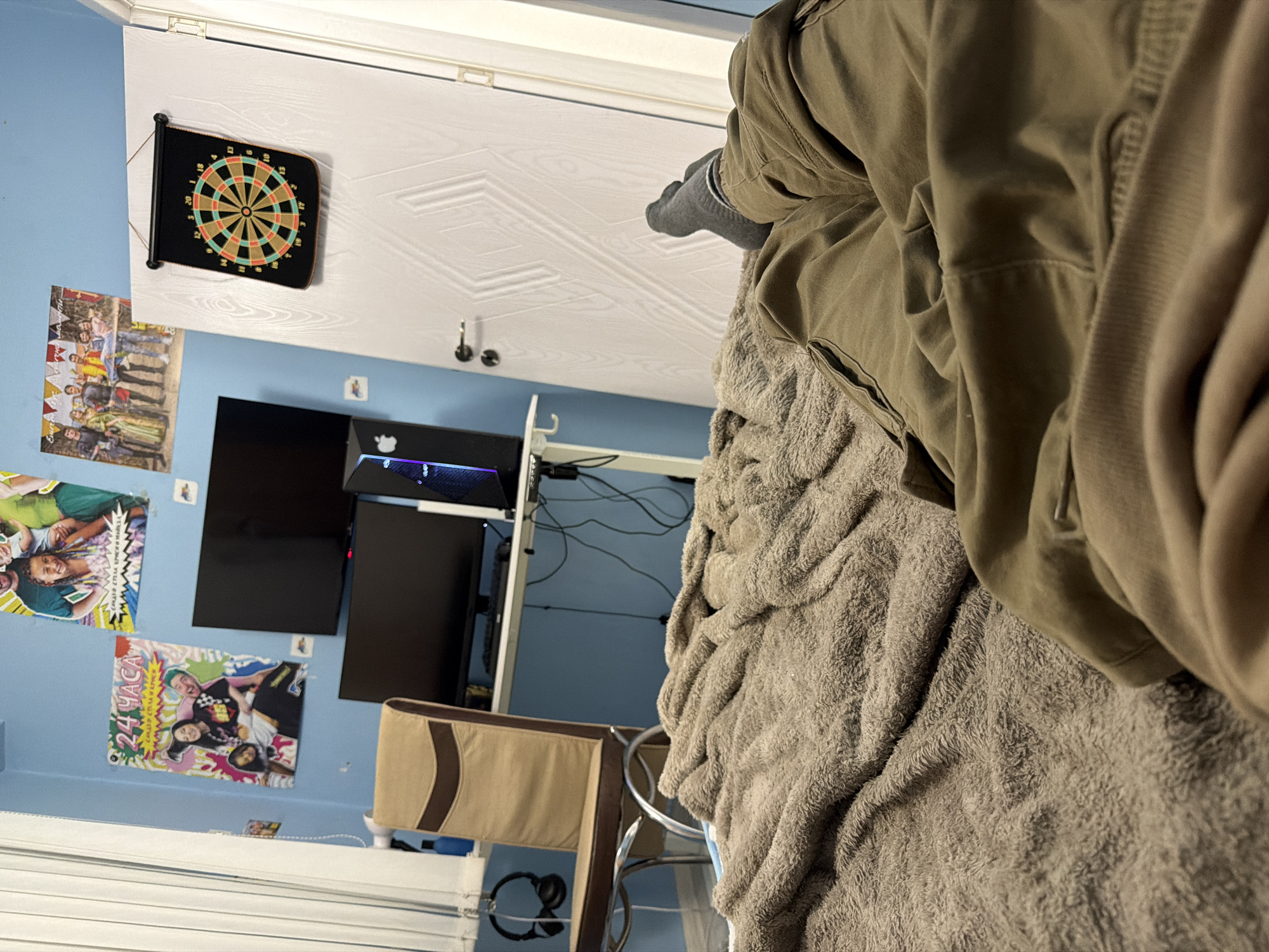 The image shows a bedroom with blue walls, a white ceiling, and a white window frame. A dartboard is mounted on the ceiling near a poster depicting people, likely a promotional image. A dual monitor setup and gaming PC are visible against the wall, alongside additional posters. A chair with a tan back is positioned near the PC. Foreground elements include a grey blanket and a person wearing grey socks and an olive green jacket. The specific location (Pernik, Bulgaria) cannot be determined from the visual information provided.