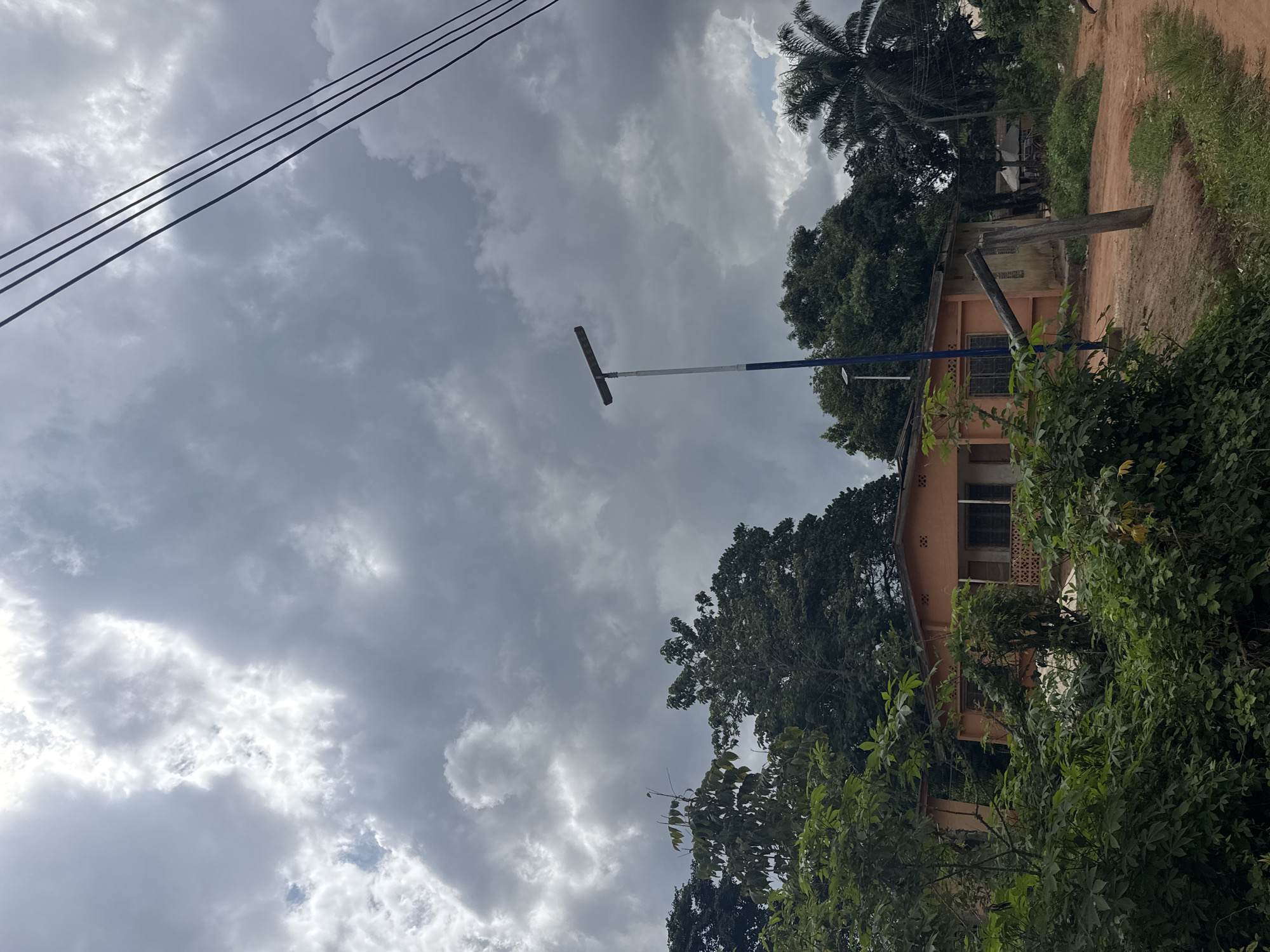 The image, likely taken in Eziudo, Nigeria, shows a building, two stories high, and tan-colored, partially obscured by trees and vegetation. A tall metal pole with a perpendicular T-shaped piece at the top extends from the ground near the building. Power lines are visible in the upper left corner of the frame, extending diagonally across the cloudy sky. To the right of the building, a dirt road is visible, bordered by more vegetation. A palm tree stands behind the building. The sky is overcast with heavy, gray clouds.