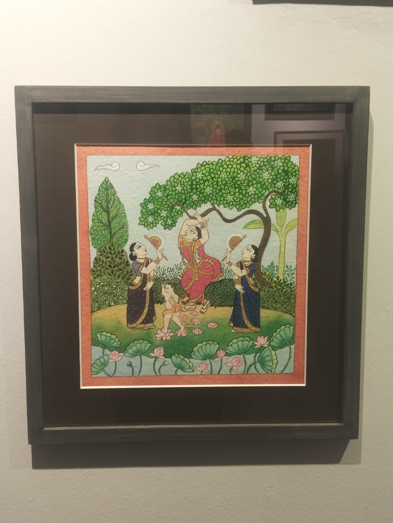 A framed painting depicting a scene with figures and nature hangs on a wall. The painting shows a figure in a red dress under a tree, with two women on either side holding fans. A child is also present, depicted standing amidst lotus flowers. The background features trees, foliage, and a light blue sky. The frame is a dark gray color. The painting is likely in an art gallery or home in Dhaka, Bangladesh, though no evidence within the image directly confirms this.