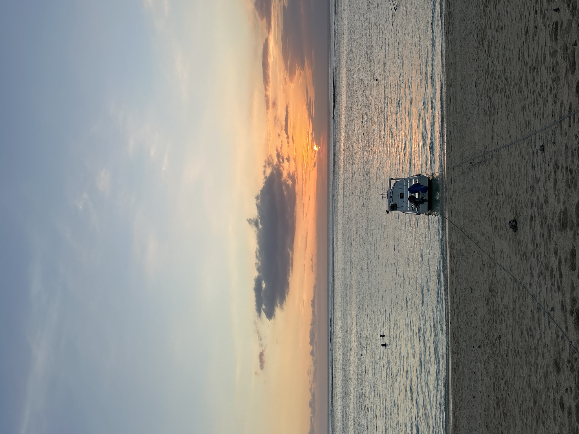 A wide-angle, vertical shot captures a serene beach scene at sunset on Nusa Penida, Indonesia. The sky displays a gradient of soft blues fading into warm oranges and pinks near the horizon, with scattered clouds tinged by the setting sun. The ocean is calm, with gentle ripples reflecting the sky's colors. In the foreground, a sandy beach stretches across the bottom of the frame, marked by the footprints of beachgoers and a few small stones. Two ropes extend from the water's edge towards the beach, anchoring a white boat that sits partially on the sand. The boat has a dark blue cushion visible on its deck and two figures, possibly preparing for departure or disembarking. Further out in the water, two individuals are visible swimming, their heads breaking the surface. The overall impression is one of tranquility and natural beauty.