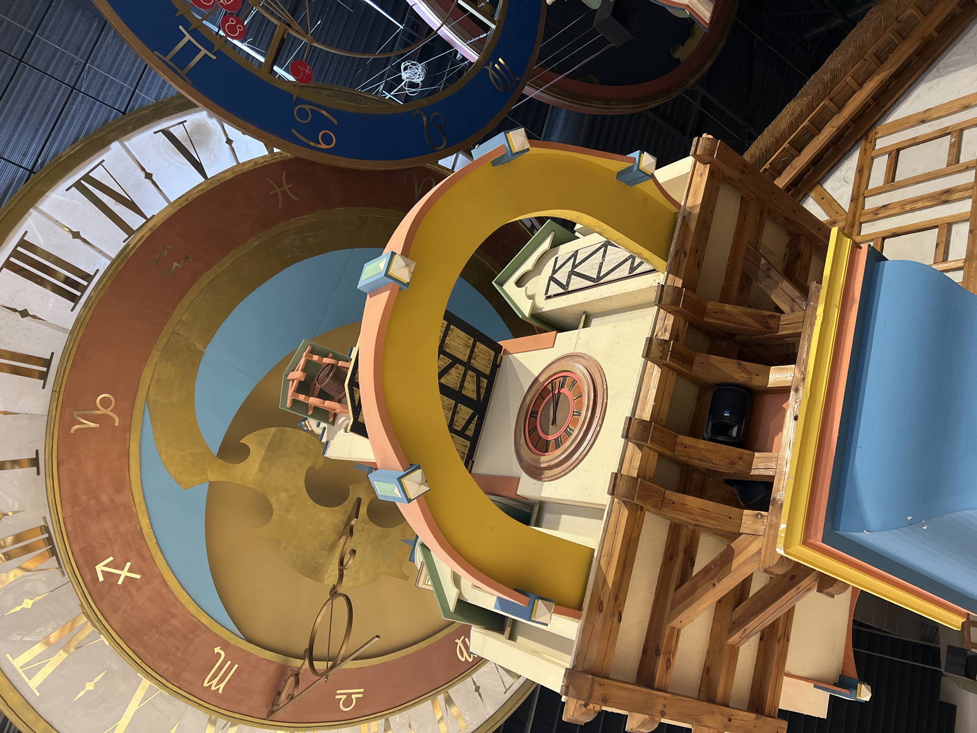 The image shows a low-angle, worm's-eye view of a section of the Klaipėda Clock Tower. The tower, adorned with vibrant colors and zodiac symbols, is centrally located in the frame. The clock tower is part of the "Annchen von Tharau" monument, located in the Old Town of Klaipėda, Lithuania, on Theater Square. The clock face itself is large and circular, featuring Roman numerals and astrological signs. Below the main clock face, the tower's architecture is characterized by wooden beams, colorful decorative elements, and smaller architectural details. The background consists of the interior of a building or dome, with visible structural elements and lighting. There are no people visible in the image.