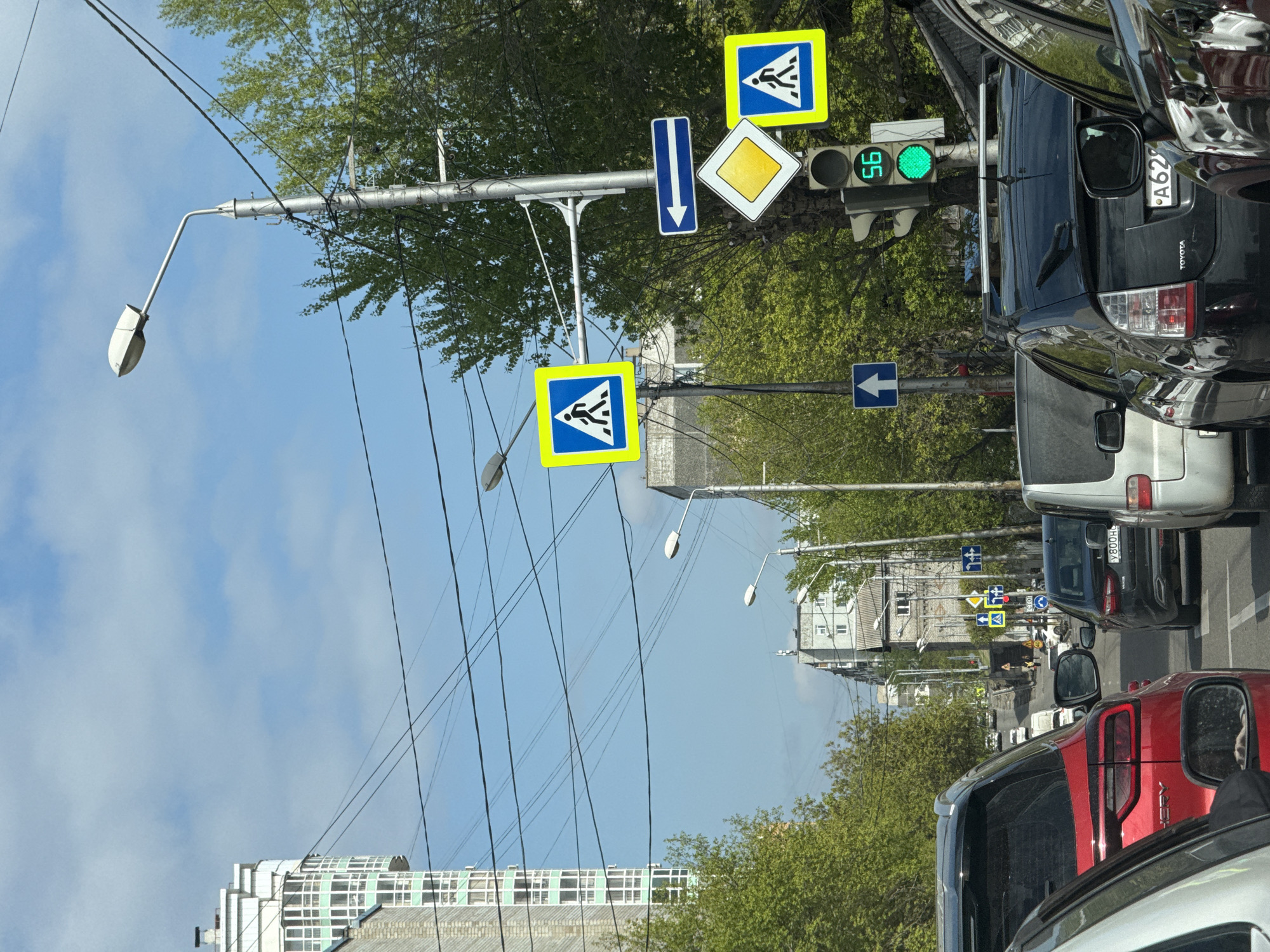 The image shows a street scene in Krasnoyarsk, Russia. The perspective is from inside a car or vehicle, likely stopped in traffic. Several vehicles are visible, including a red SUV and a black Toyota Prius. Street signs indicating pedestrian crossings, a one-way street, and a yield sign are seen. A traffic light is illuminated in green. The backdrop includes trees, buildings, and a sky with clouds. Utility poles and wires are also visible.