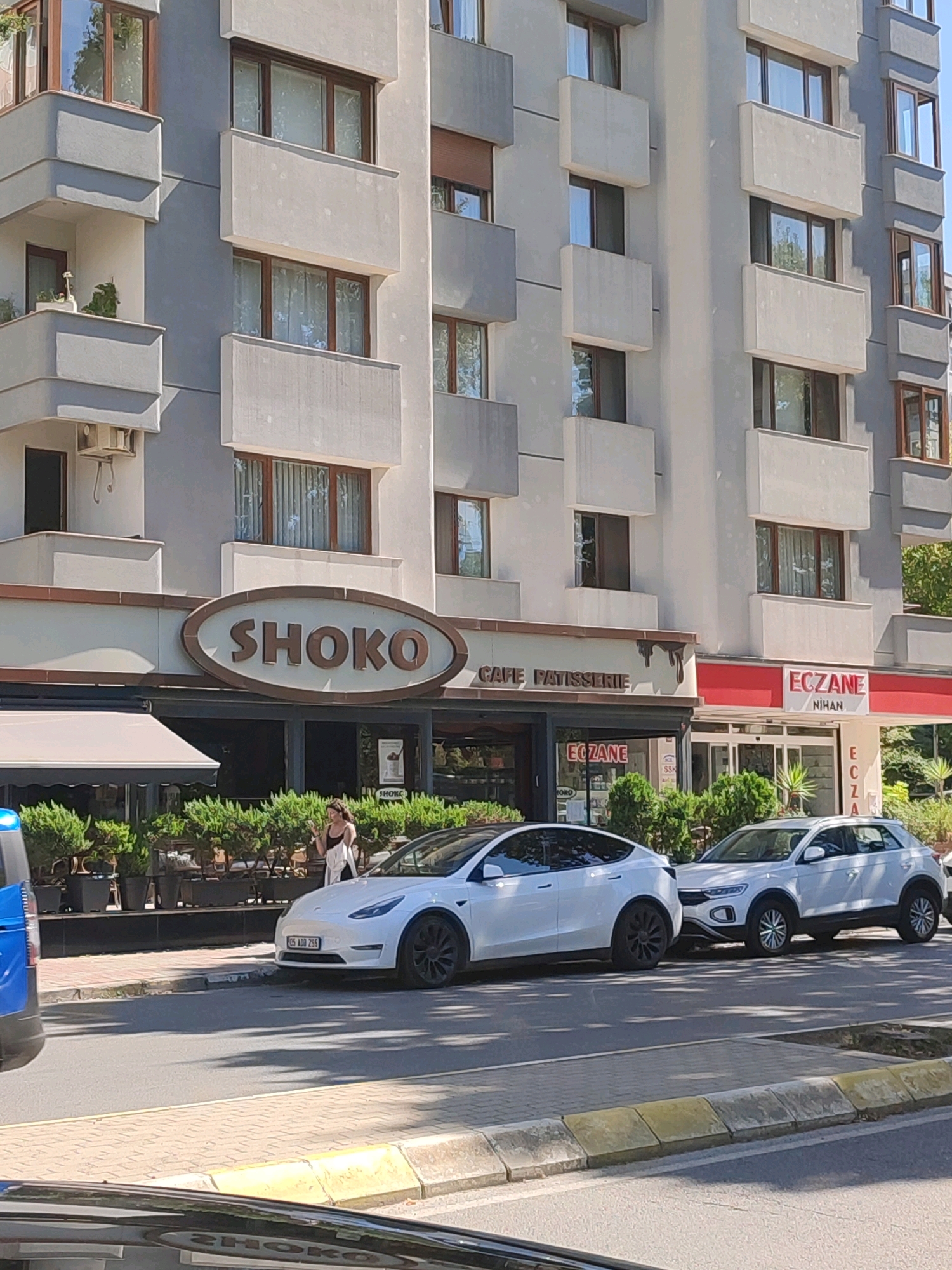 This is a medium shot, eye-level, street-level photograph taken in Kadıköy, Turkey. The scene depicts a bright, sunny day with a clear blue sky. In the foreground, a white Tesla Model Y is parked on the street, facing left. To its right, a white Volkswagen T-Roc is also parked. A blue and white vehicle, possibly a police car, is partially visible on the far left.
Behind the cars, a row of businesses lines the sidewalk. A prominent sign with the word "SHOKO" in large, brown letters on a brown oval background is centered above the entrance to a cafe and patisserie. To the right of the "SHOKO" establishment, there is a pharmacy, identified by a red and white sign that reads "ECZANE" with "NİHAN" below it. The cafe has a large glass storefront with greenery visible inside and outside, and an awning extending over the sidewalk.
A woman wearing a white top and dark bottoms is standing on the sidewalk near the cafe entrance, looking at her phone. She is positioned between the cafe and the pharmacy. The buildings in the background are multi-story residential and commercial structures with balconies and windows. Trees and planters with manicured bushes are situated along the sidewalk in front of the businesses. The street is paved and shows some wear. The lighting suggests it is daytime, with shadows cast on the street and sidewalk.
