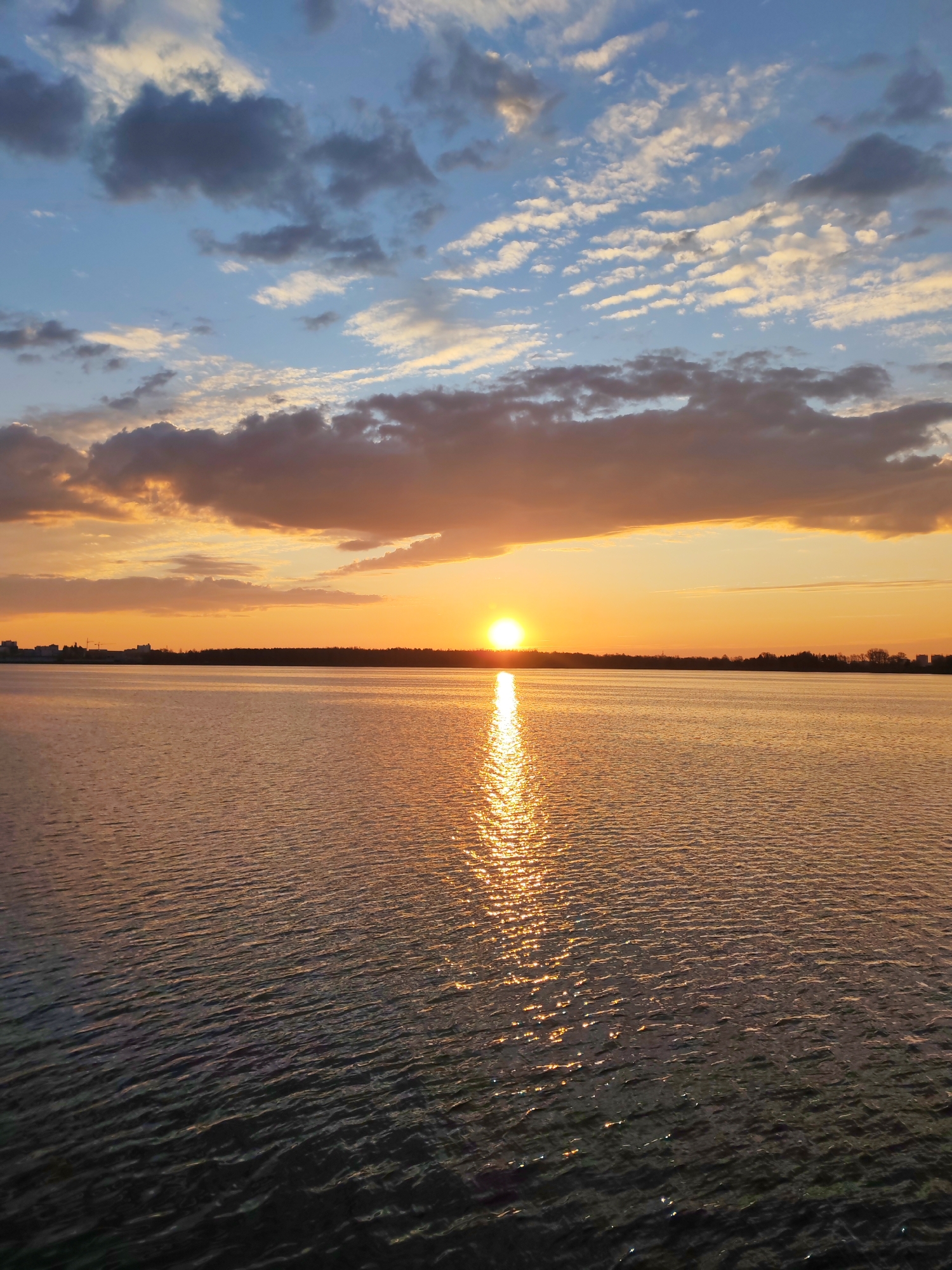 The image captures a sunset over a body of water. The sun is positioned near the horizon, creating a bright reflection on the water's surface. The sky is a mix of blues and oranges, with clouds scattered throughout. In the distance, there is a dark strip of land that appears to be a treeline or a cityscape. The water has small ripples across it. The location is Unknown, Unknown.