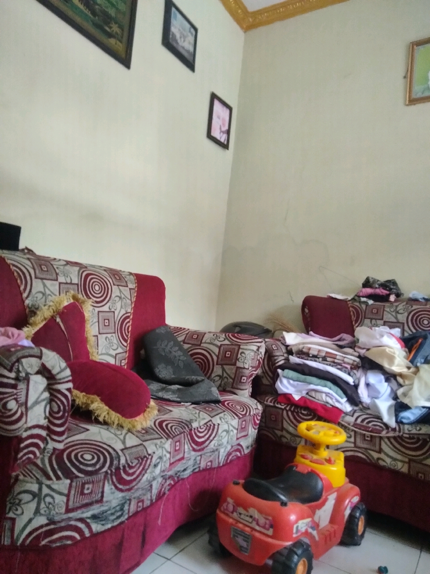 The image depicts an indoor scene, likely a living room, in a residence in Suka Sari, Indonesia. Two red sofas with patterned upholstery are visible. The sofa on the left has decorative pillows, including a red one with gold fringe. The sofa on the right has a pile of folded clothing on it. In front of the right sofa is a red and yellow toy car. The walls are a pale color, and there are several framed pictures hanging on them. One picture on the left wall appears to be a landscape painting, and two smaller framed pictures are on the right wall. The overall impression is of a moderately furnished and lived-in space.