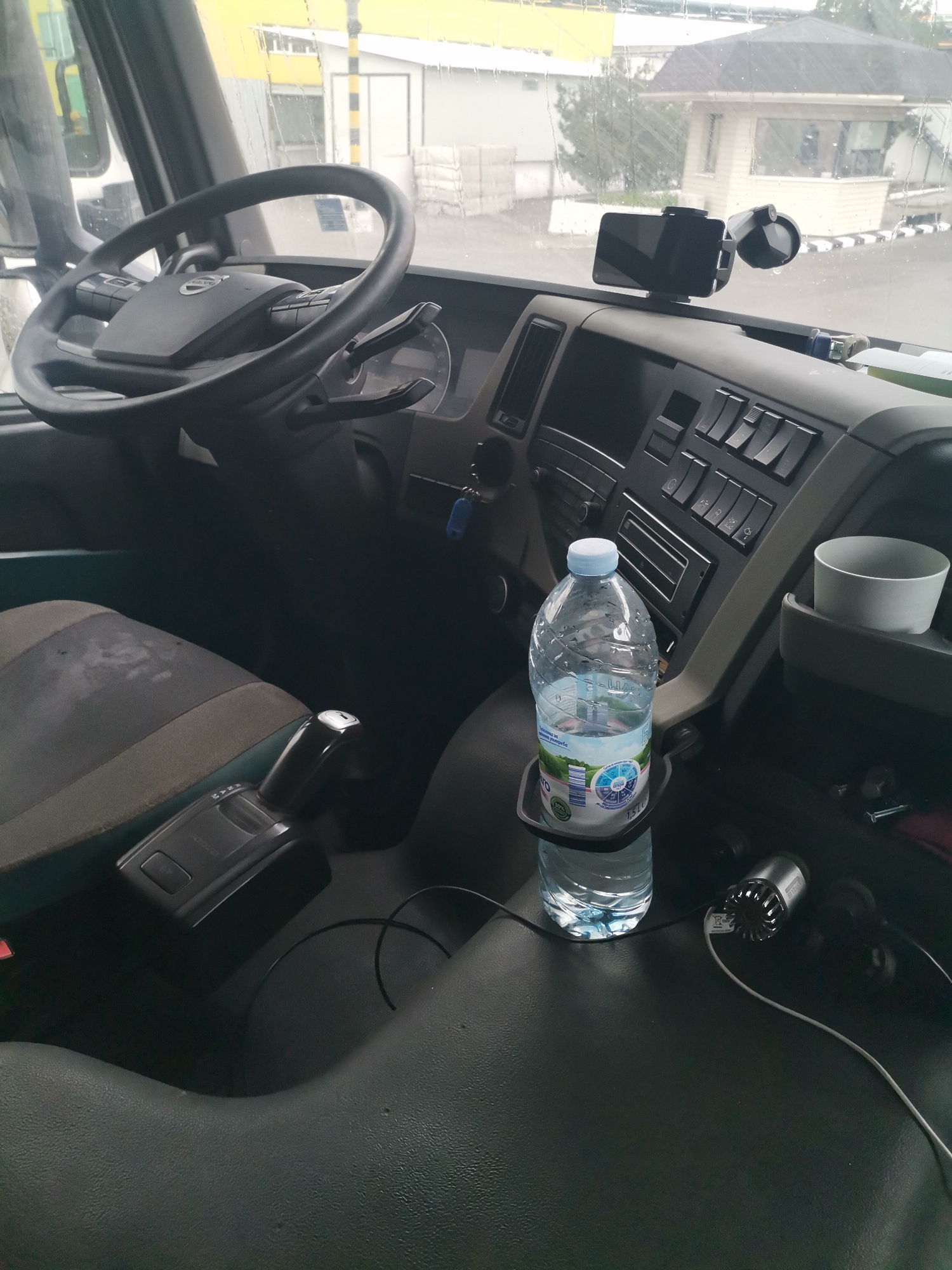 The image provides a view from the interior of a vehicle, specifically the cabin of a Volvo truck, looking forward through the windshield. The steering wheel, featuring the "Volvo" logo, dominates the left side of the frame.
Through the rain-streaked windshield, an exterior scene unfolds, showing several buildings, including a small, light-colored guardhouse or office with a dark roof and a striped barrier, indicative of an entrance to an industrial or commercial facility. The setting is Karapelit, Bulgaria, suggesting the truck is parked or waiting at such a location.
On the dashboard, a smartphone is secured in a mount near the windshield. To the right, a white ceramic cup sits in a built-in holder. A 1.5-liter plastic water bottle, labeled with "ВОДА" (VODA - Water) and "PIRIN" (or similar, partially visible Cyrillic text), is placed in a cup holder between the driver and passenger seats. Below the water bottle, a small electronic device with a fan-like grille is connected by wires, possibly a charger or power inverter. Keys are visible hanging from the ignition.
The driver's seat, upholstered in a combination of grey and green fabric, shows signs of wear and some stains. The gear selector, located to the right of the driver's seat, has text that includes "AUTORIVE" (likely "AUTO" and indicating drive modes). The dashboard is equipped with numerous rocker switches and controls.
The overall impression is of a working truck, stationary during the daytime, potentially experiencing or having recently experienced rain, at what appears to be a facility gate.