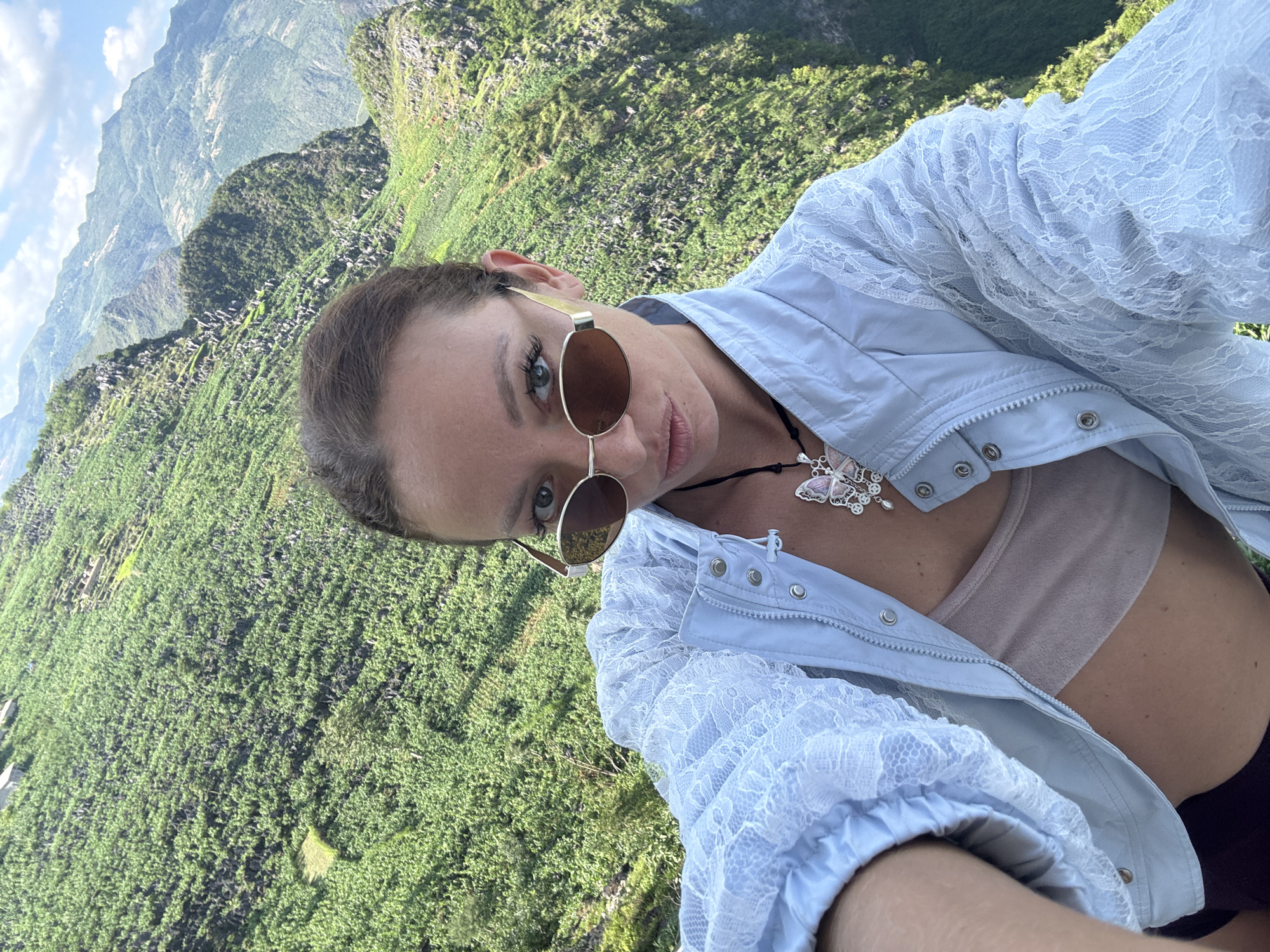 A woman wearing sunglasses and a light blue lace-trimmed jacket is pictured in a selfie. The background reveals a mountainous landscape covered in dense green vegetation. Based on the landscape and presence of terraced fields, the location is likely in the Đồng Văn Commune, Vietnam.