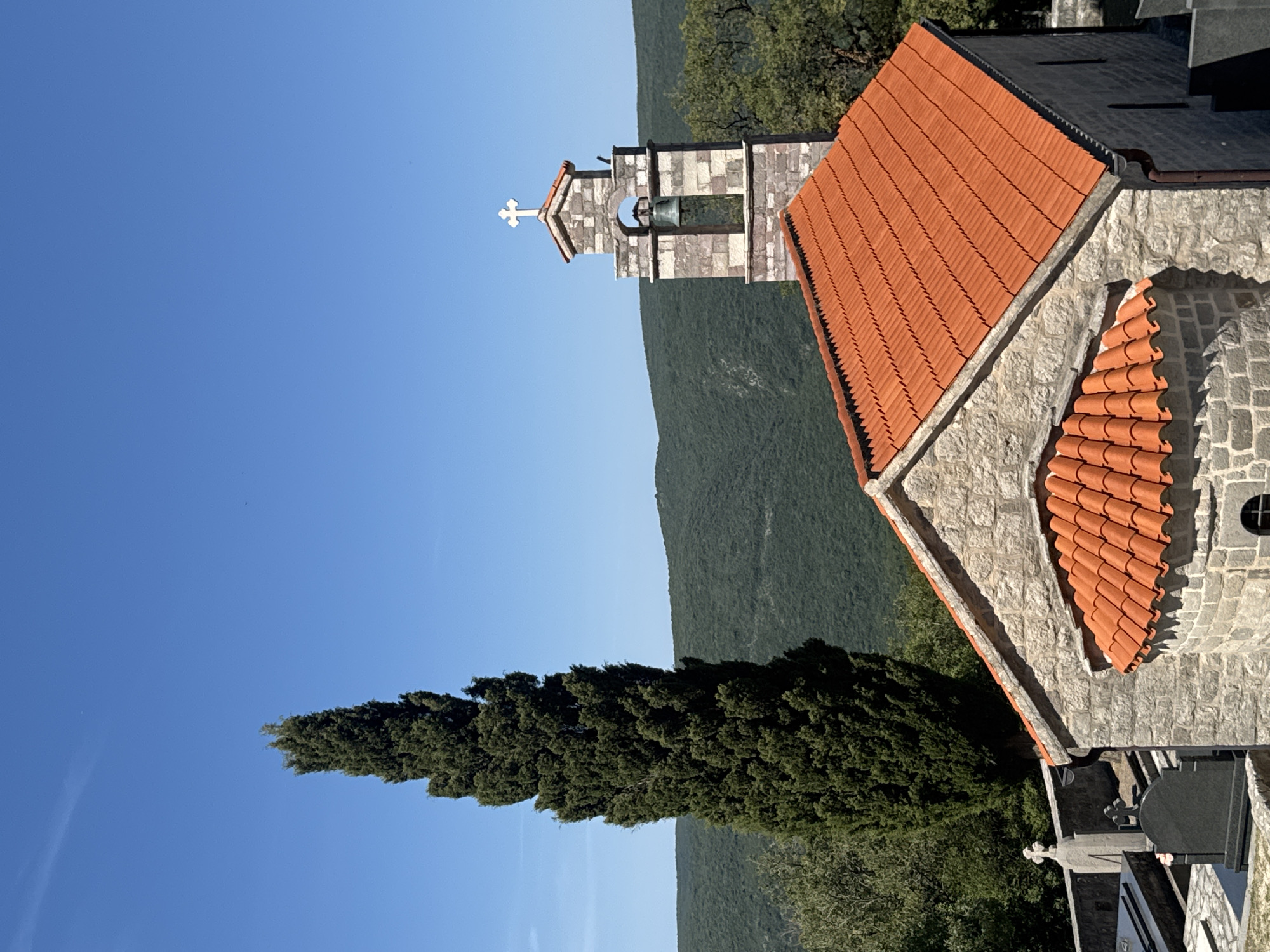 The image depicts a stone church with an orange tiled roof and a bell tower topped with a cross, set against a backdrop of rolling green hills under a clear blue sky. In the foreground, to the left of the church, are gravestones, indicating a cemetery. A tall, slender cypress tree stands to the left of the bell tower. The church appears to be situated in a rural or semi-rural setting, likely within the village of Bratešići, Montenegro, given the context provided. The scene is peaceful and serene, bathed in natural daylight. No people or animals are visible in the image.