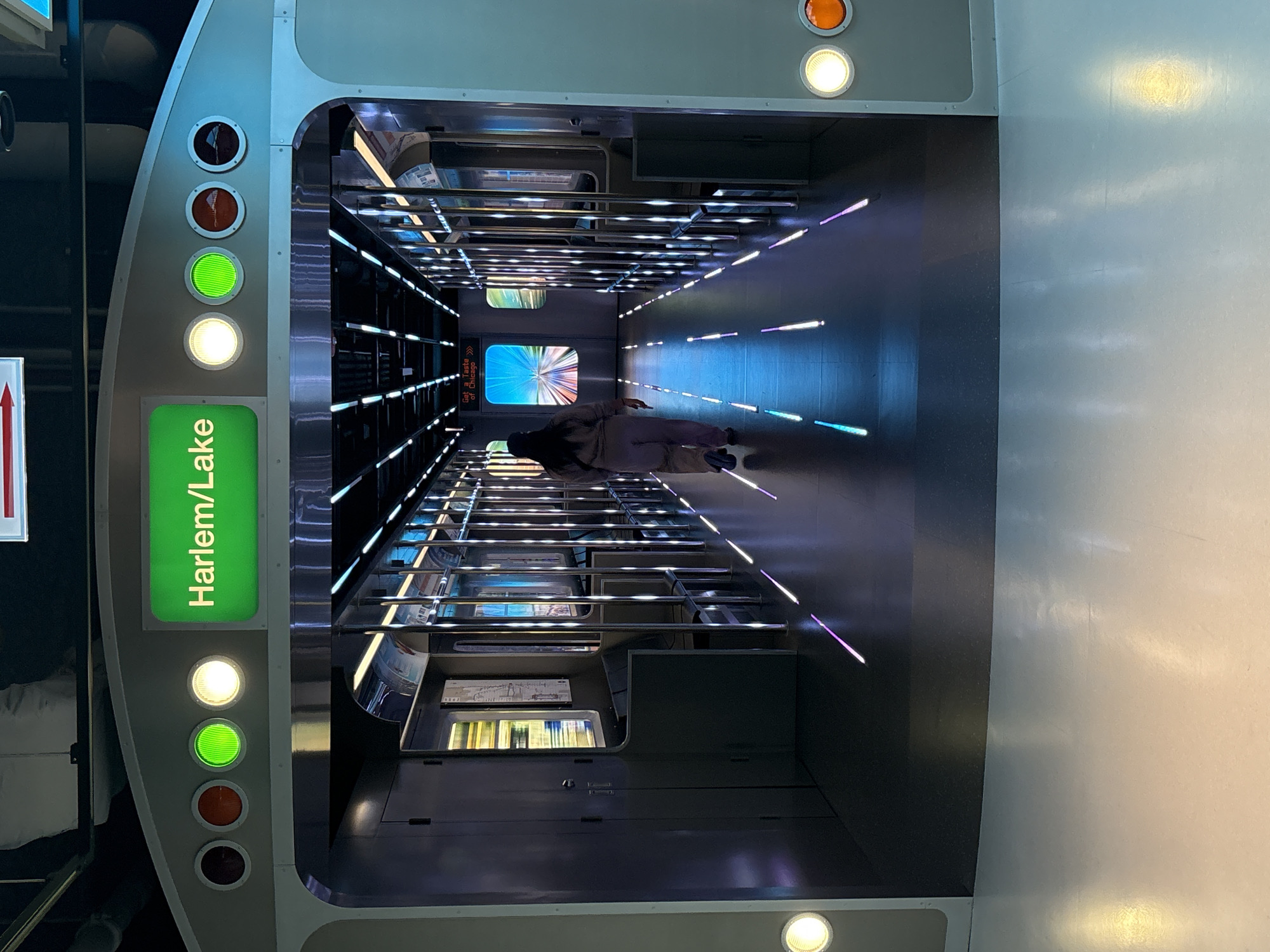 The image shows an exhibit of a train car, featuring the "Harlem/Lake" sign, likely representing the Harlem/Lake station on the Chicago Transit Authority (CTA) Green Line. The car's interior is illuminated with streaks of light on the floor. A person lies on the floor of the car.