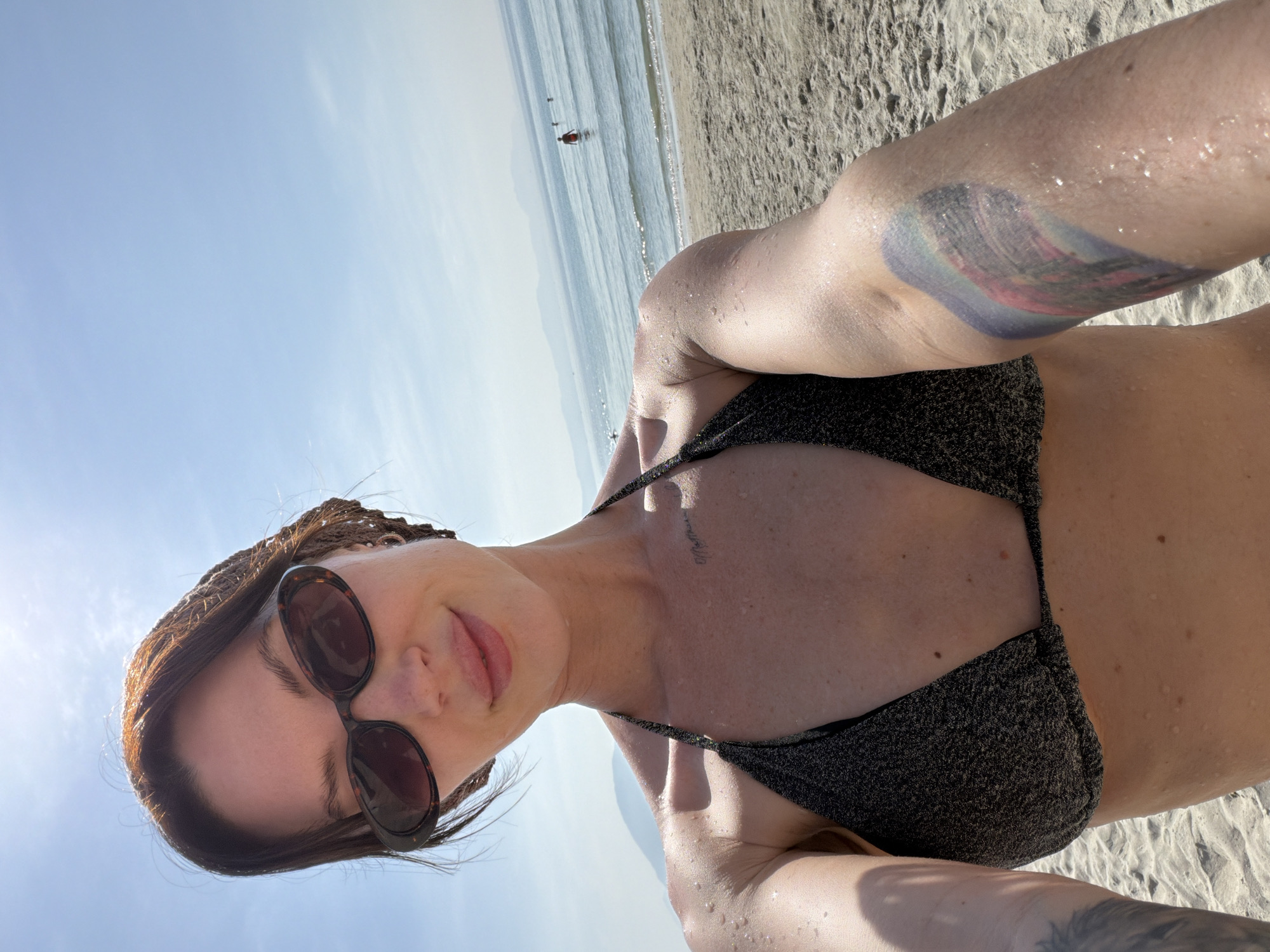 The image presents a close-up selfie of a female individual on a beach in Dong Ninh Hoa Ward, Vietnam. The woman, appearing to be between 20 and 40 years old, has dark hair partially covered by a brown woven headscarf. She wears dark, oval-shaped sunglasses and a dark, glitter-textured bikini top. Numerous water droplets are visible on her exposed skin, particularly on her arms and chest. A small text tattoo is located on her upper chest, and a larger, colorful, abstract tattoo, featuring hues of blue, purple, and red, is present on her left upper arm. She displays a slight smile.
The background depicts a sandy beach adjacent to a calm, light-blue ocean under a clear, bright blue sky. Several indistinct figures are visible in the ocean, with one individual wearing a red garment wading in the mid-ground. Hazy mountains are discernible on the distant horizon line. The scene is brightly illuminated by high sunlight.