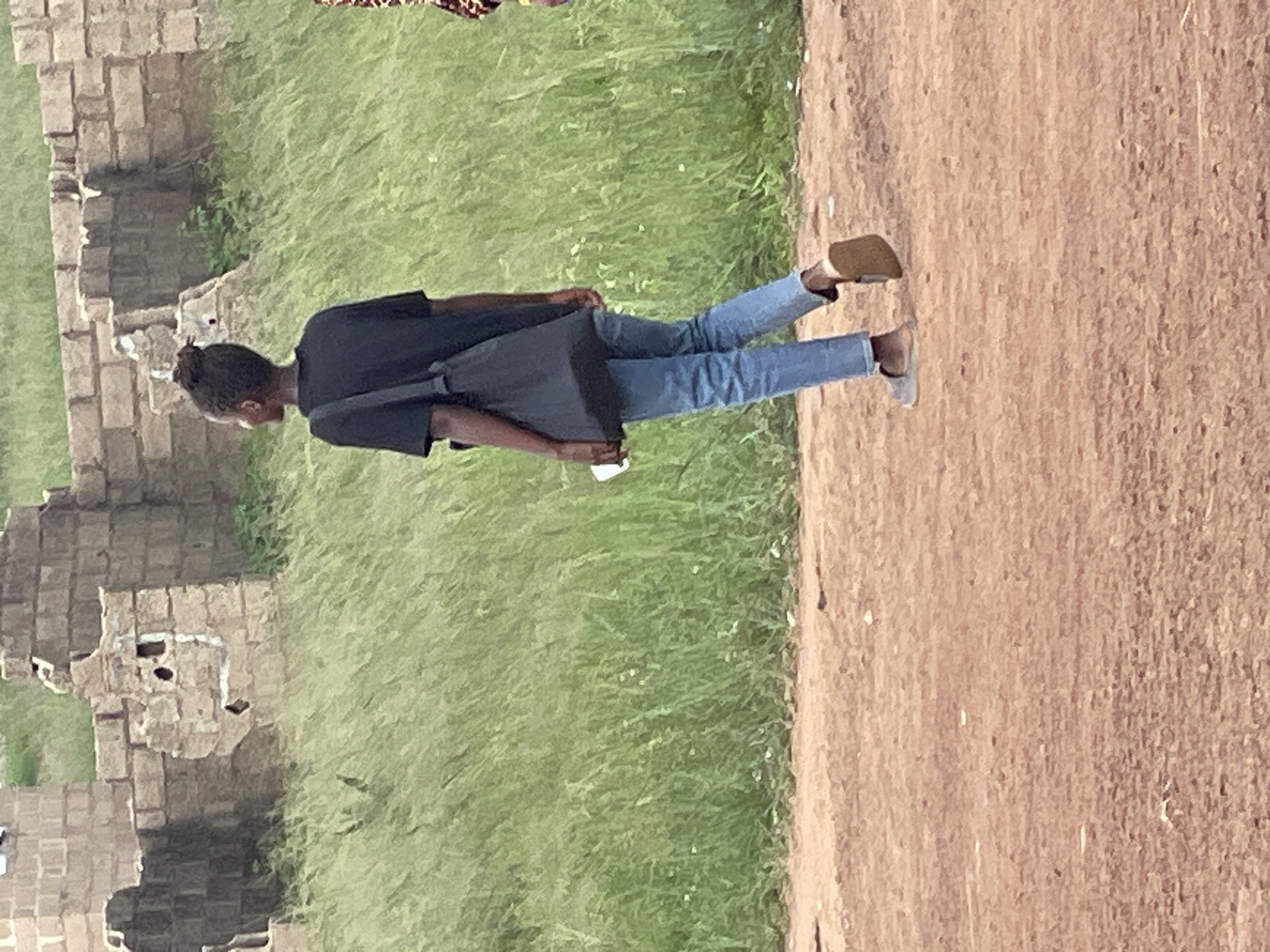 A young Black person with dark hair styled in a bun walks away from the camera on a dirt path. The person is wearing a black t-shirt, blue jeans, and light-colored sandals. A black messenger bag is slung over their left shoulder, and they are holding a white object in their right hand. In the background, there are overgrown green grasses and the remains of a stone building with rough-hewn blocks. The scene appears to be in Durumi, Nigeria.