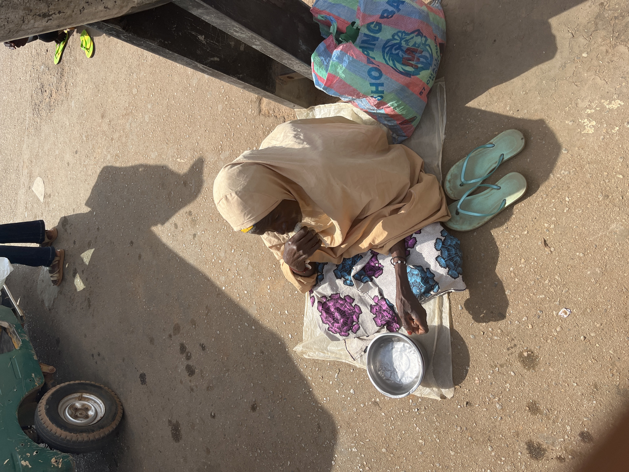 An elderly woman wearing a light brown headscarf and a colorful patterned dress is sitting on the ground in Gura Top, Nigeria. She is on a white plastic sheet next to a metal bowl. Her hands are raised to her mouth as she appears to be eating. Beside her are a pair of light blue flip-flops and a large, striped shopping bag. A green vehicle is partially visible to the left of the frame, and a dark wooden structure is to the right. The ground is a mix of sand and dirt, with shadows cast by the sun. In the background, two pairs of legs are visible, one wearing dark pants and sandals, and the other wearing bright yellow flip-flops.