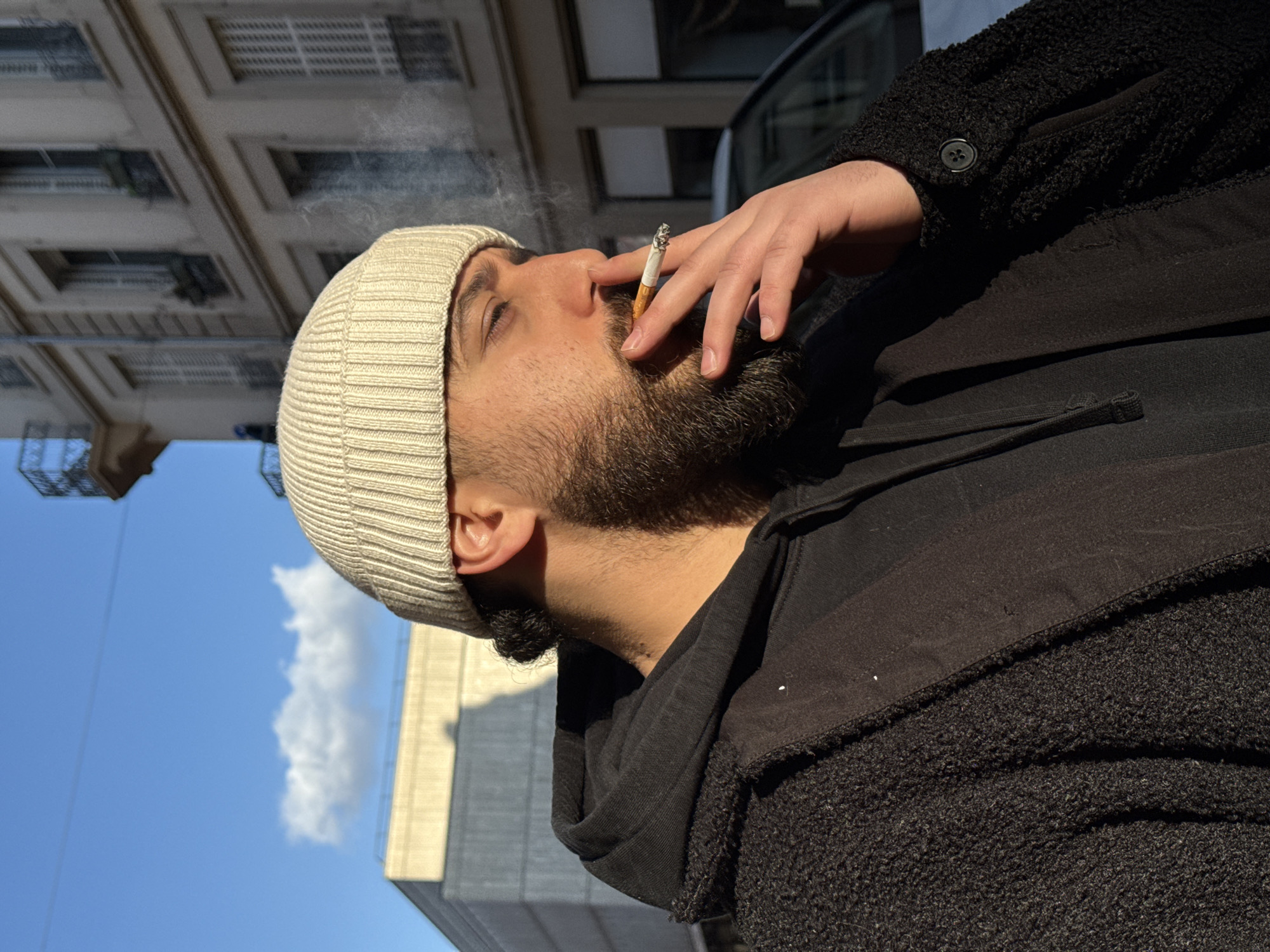 The image presents an urban outdoor scene under a clear blue sky with scattered white clouds. A male subject is depicted in a mid-shot, angled towards his right, with direct sunlight illuminating the right side of his face and body. He wears a light-colored ribbed beanie, a black hooded sweatshirt, and a dark, textured open coat. The subject is holding a lit cigarette between the index and middle fingers of his right hand, positioned at his mouth, with visible smoke rising from it. In the background, a modern building with a flat roof is partially visible to the left. To the right, a multi-story classical European building with light-colored walls, numerous windows featuring closed white shutters, and small balconies with railings is present. A light-colored vehicle is partially discernible behind the subject's right arm. The architectural characteristics of the background buildings indicate an urban location within Strasbourg, France.