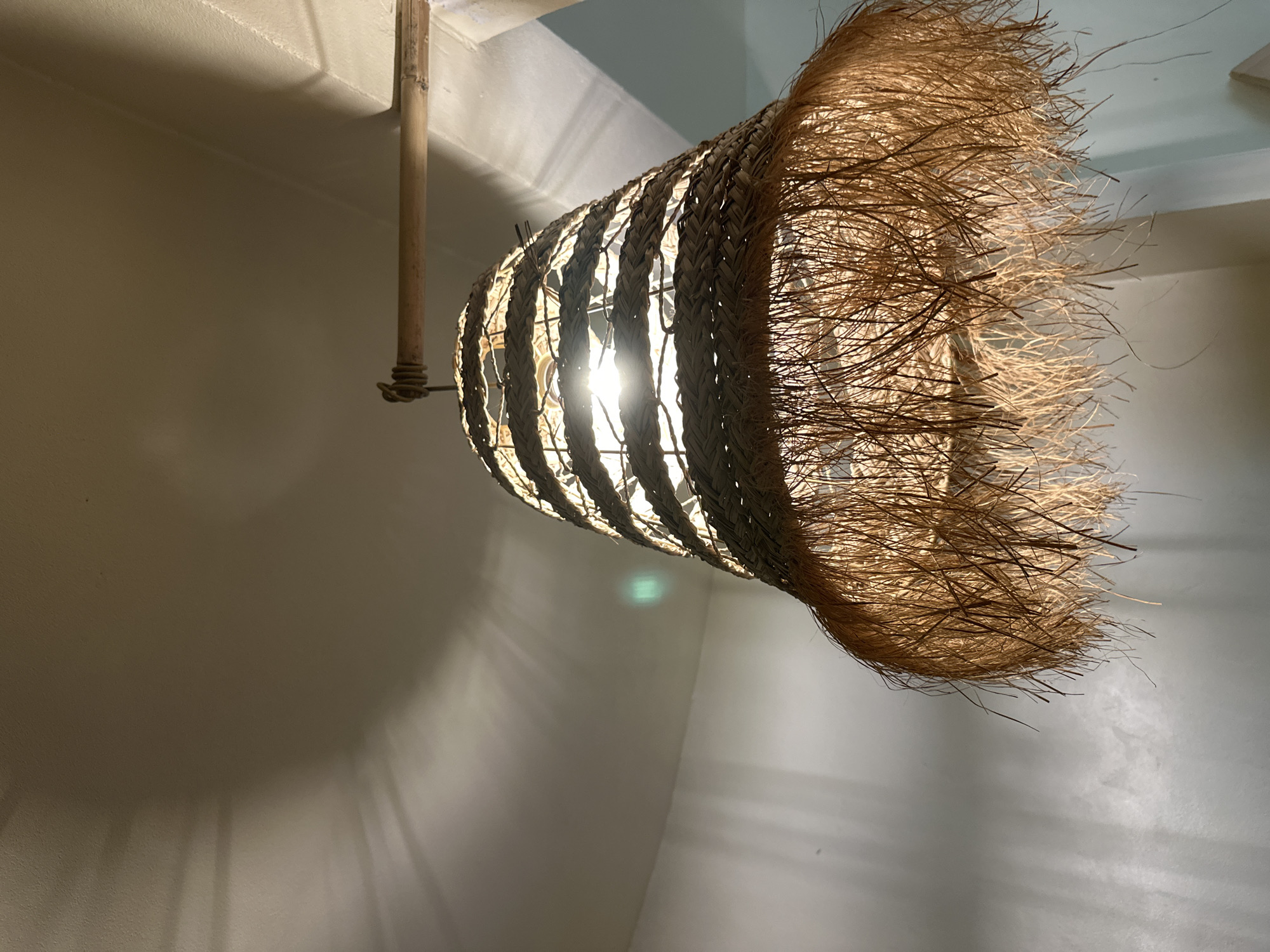 The image presents an upward view of an illuminated light fixture, likely a pendant lamp, hanging in an indoor setting. The central subject is a distinct lampshade crafted from natural, light-brown fibers, possibly straw, rattan, or similar woven material. The top section of the lampshade features a more structured, braided or woven pattern, through which the bright light source within is visible, casting a strong glow and distinct light rays. The lower portion of the lampshade flares out into a thick, shaggy fringe of fine, dry strands, creating a unique, organic, and somewhat wild texture.
The light fixture is suspended from the ceiling by a simple mechanism that includes a natural wooden pole, possibly bamboo, visible on the left side of the frame. The strong light from the lamp casts intricate shadows across the surrounding walls and ceiling, which are painted in a light, neutral tone, appearing off-white or light beige. A soft, greenish luminescence is noticeable on a wall surface in the background, possibly a reflection or ambient light.
The scene suggests an interior space, where artificial light is the primary illumination. The natural materials of the lamp and its design evoke a rustic, bohemian, or tropical aesthetic, consistent with the location context of ตำบลเกาะพะงัน, Thailand. The overall impression is one of warmth and an inviting ambiance, with the light creating a cozy and decorative effect. No visible text is present in the image.
