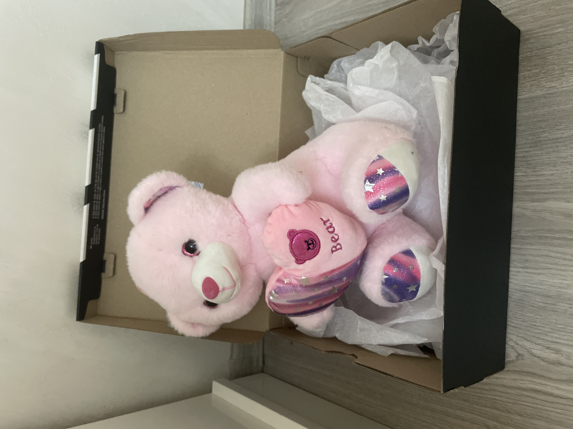 An overhead, slightly tilted shot captures a pink teddy bear nestled within an open cardboard box, likely indoors in Ihtiman, Bulgaria. The teddy bear, made of soft pink plush, has bright, sparkly eyes and a lighter pink snout with a darker pink nose. It is holding a smaller, darker pink heart-shaped pillow to its chest, which features the embroidered word "Bear" and a small pink bear head icon. The bear's hands and feet have distinctive iridescent fabric inserts with a pattern of horizontal pink, purple, and blue stripes adorned with small white stars.
The bear rests on crinkled white tissue paper inside a brown cardboard box. The right side of the box is black, suggesting it might be an outer sleeve or a black lid component. The left inner edge of the box is visible, showing thin black and white stripes with some faint, illegible text or markings.
The box is placed on a flat, hard white surface, possibly a shelf or table. A plain white wall forms the background on the left, and a light grey wooden floor or surface is visible on the right and bottom of the frame. There is no discernible activity beyond the static arrangement of the bear in the box, suggesting it is either packed, unpacked, or stored. No people are visible. The lighting suggests an indoor setting, likely daylight or bright artificial light.