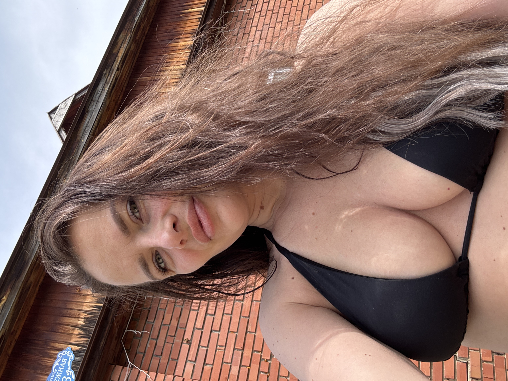 A woman with long brown hair poses outdoors, wearing a black bikini top. A brick wall is behind her. The location is Rusalevka, Russia, as indicated by a sign written in Russian on the bottom left of the image.