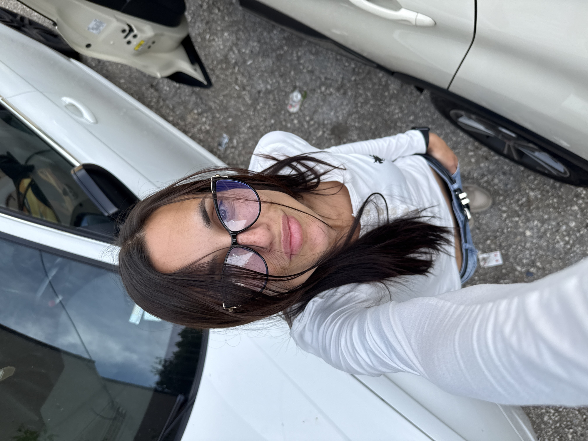 A woman is taking a selfie outside in an urban environment, likely a parking lot or street with multiple cars. The cars are mostly white. The woman has dark hair and is wearing glasses, a white long-sleeved shirt, and jeans. A discarded beverage container is visible on the ground. I cannot confirm that the location is in Pleven, Bulgaria from the provided image.