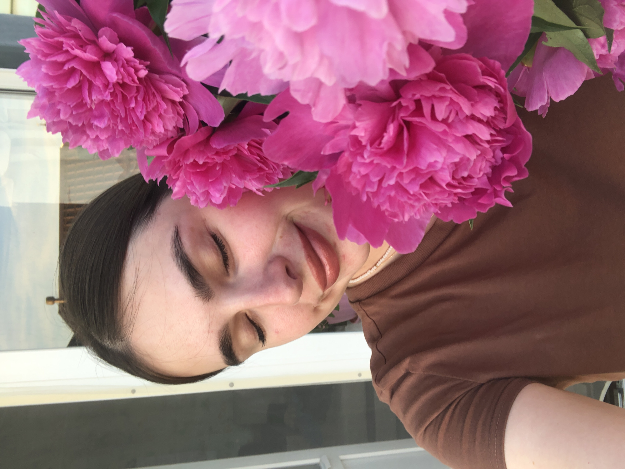 A young woman is captured in a close-up shot, her face tilted upwards amidst a bouquet of vibrant pink peonies. Her eyes are closed, and her lips are slightly parted, conveying a sense of peace or perhaps a moment of enjoyment. The soft, natural light suggests it might be daytime. She is wearing a simple brown t-shirt. The background is out of focus, but appears to be an outdoor setting with a glimpse of a window or reflective surface and what looks like a building. A small, metallic hair accessory is visible in her dark hair. The overall mood is serene and gentle, highlighting the beauty of the flowers and the woman's soft expression.