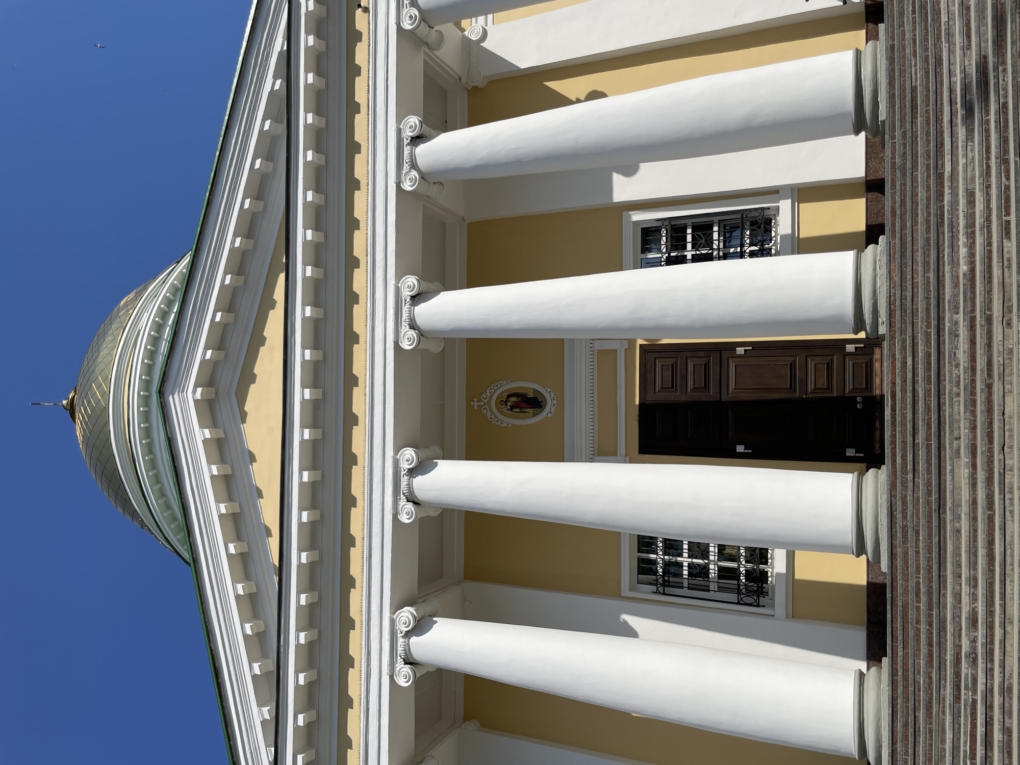The image depicts the facade of a building in Izhevsk, Russia, notable for its classical architecture. The building features a golden dome partially visible against a blue sky. The facade is characterized by a series of white columns with decorative capitals, supporting an entablature. The wall behind the columns is painted in a pale yellow color. There are two windows with decorative metal grilles and a dark wooden door. An oval-shaped decorative element is visible on the wall between the columns. To the right, there is a section of the building with a vertical striated pattern.