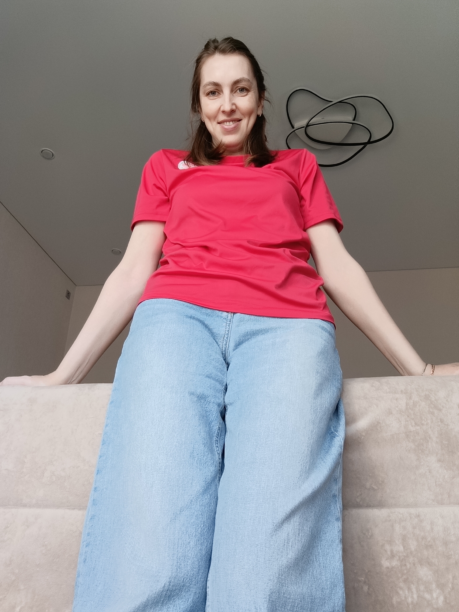 This low-angle, full-shot portrait features a smiling woman in an indoor setting, likely in Chaikovsky, Russia. The woman, appearing to be in her 30s or 40s, is positioned as if seated or perched on the backrest of a light beige sofa, looking directly at the camera with a natural, pleasant smile. She has light brown hair, which is pulled back.
She is wearing a bright red, short-sleeved t-shirt with a small white logo resembling a Nike swoosh on the left chest. Her lower body is covered by light blue, wide-leg jeans. Her arms extend outwards, resting casually on the sofa's backrest.
The background shows a modern room with light grey or off-white walls and ceiling. Visible ceiling elements include a recessed light on the left and a contemporary, abstract chandelier with intertwined black and white components on the right. The lighting is soft and even, suggesting either natural light from an unseen window or well-distributed artificial illumination, creating a relaxed and inviting atmosphere.